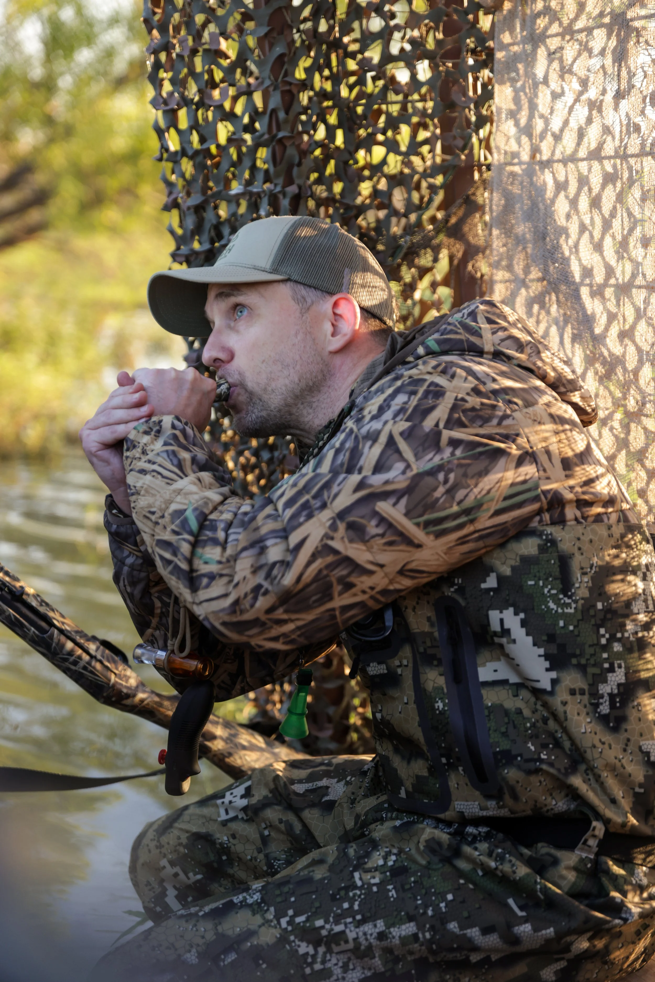 A man in camouflage clothing and a gray cap is sitting near water, hunting or observing wildlife, with a provided camouflage curtain or net behind him, during the daytime.