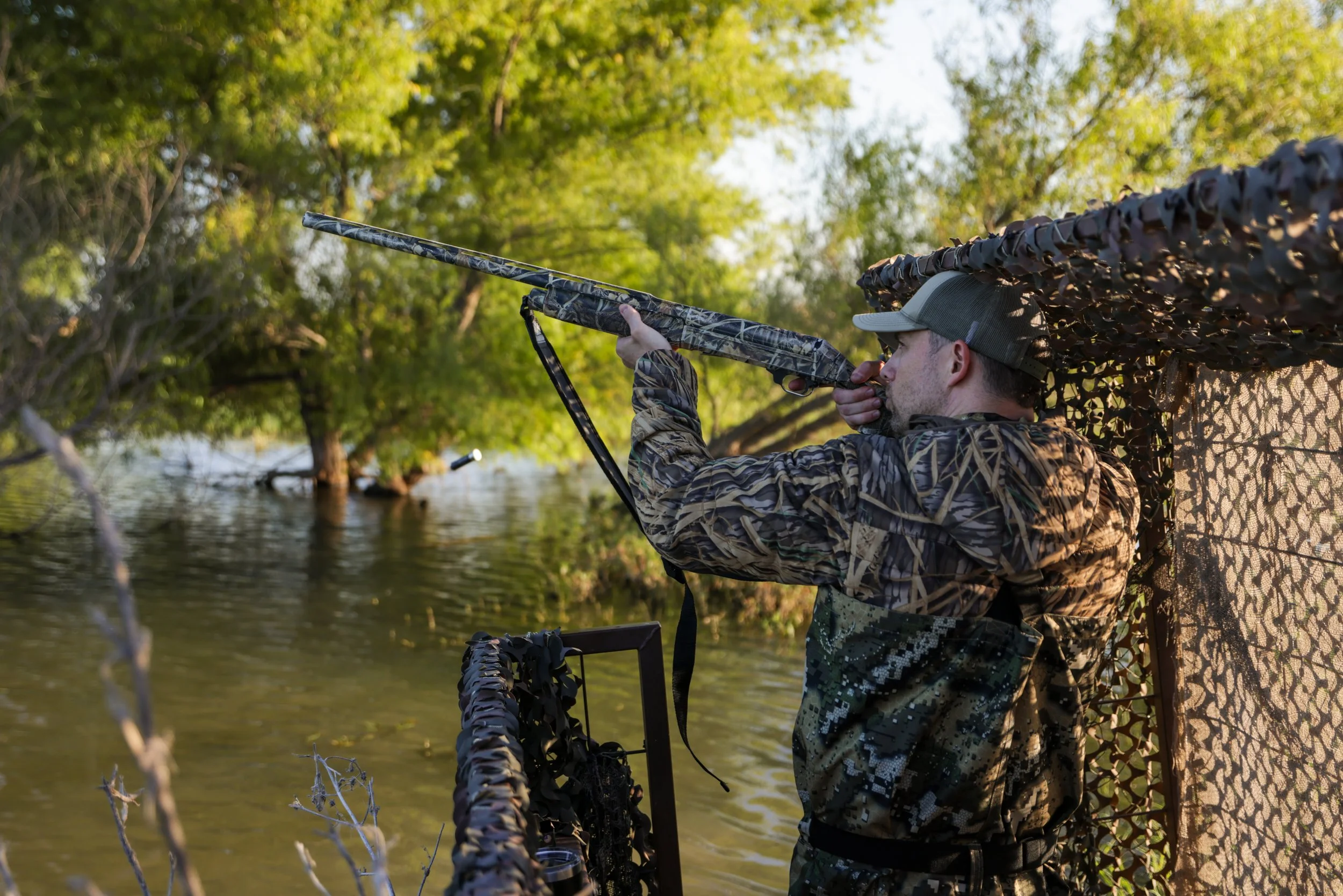 A man in camouflage clothing and a hat aims a camouflage rifle from behind a camouflage hunting blind near a body of water surrounded by trees.