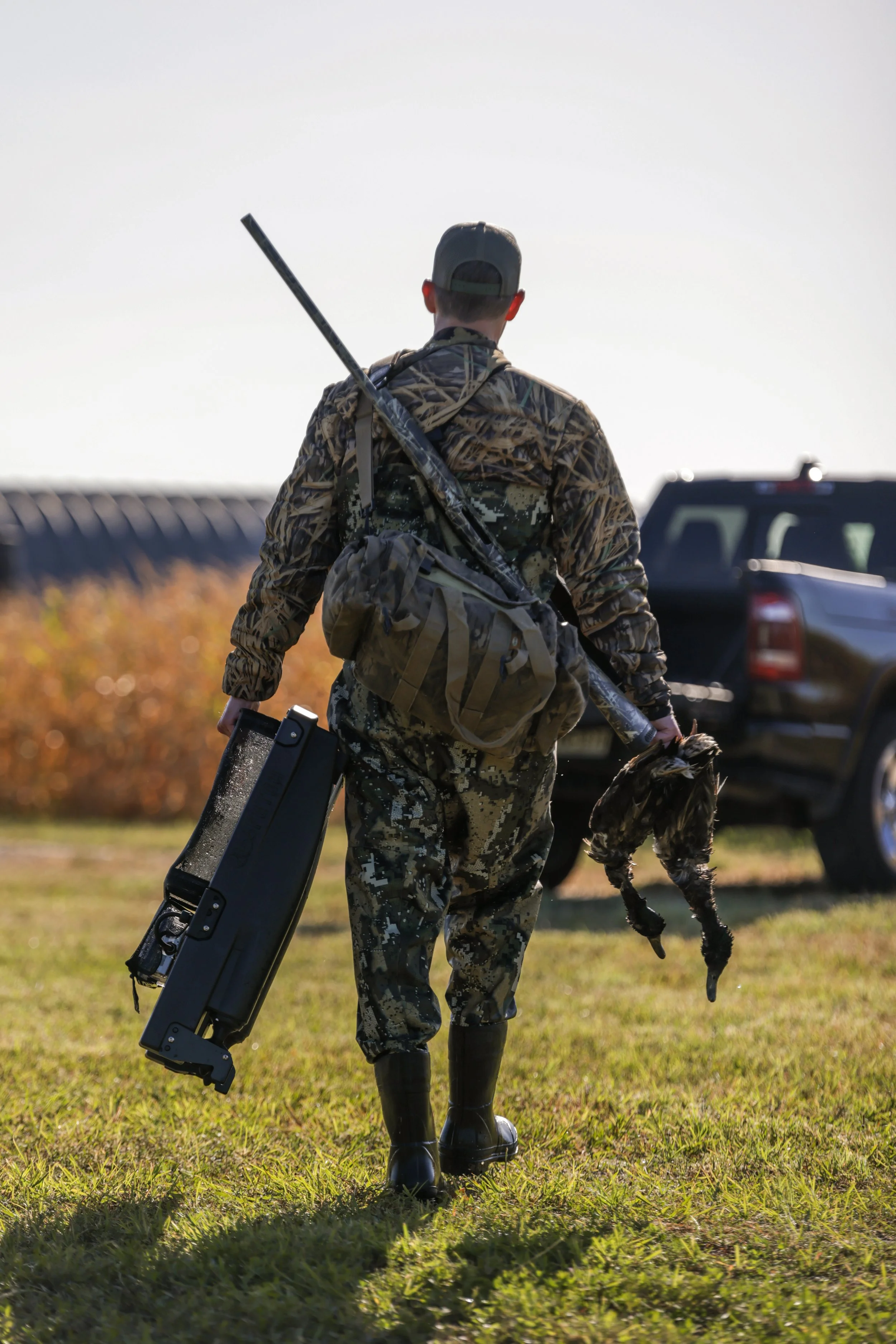 A person in camouflage hunting gear is carrying a rifle and a Fetch waterfowl retrieval system while standing next to a black pickup truck in a field with tall cornstalks.