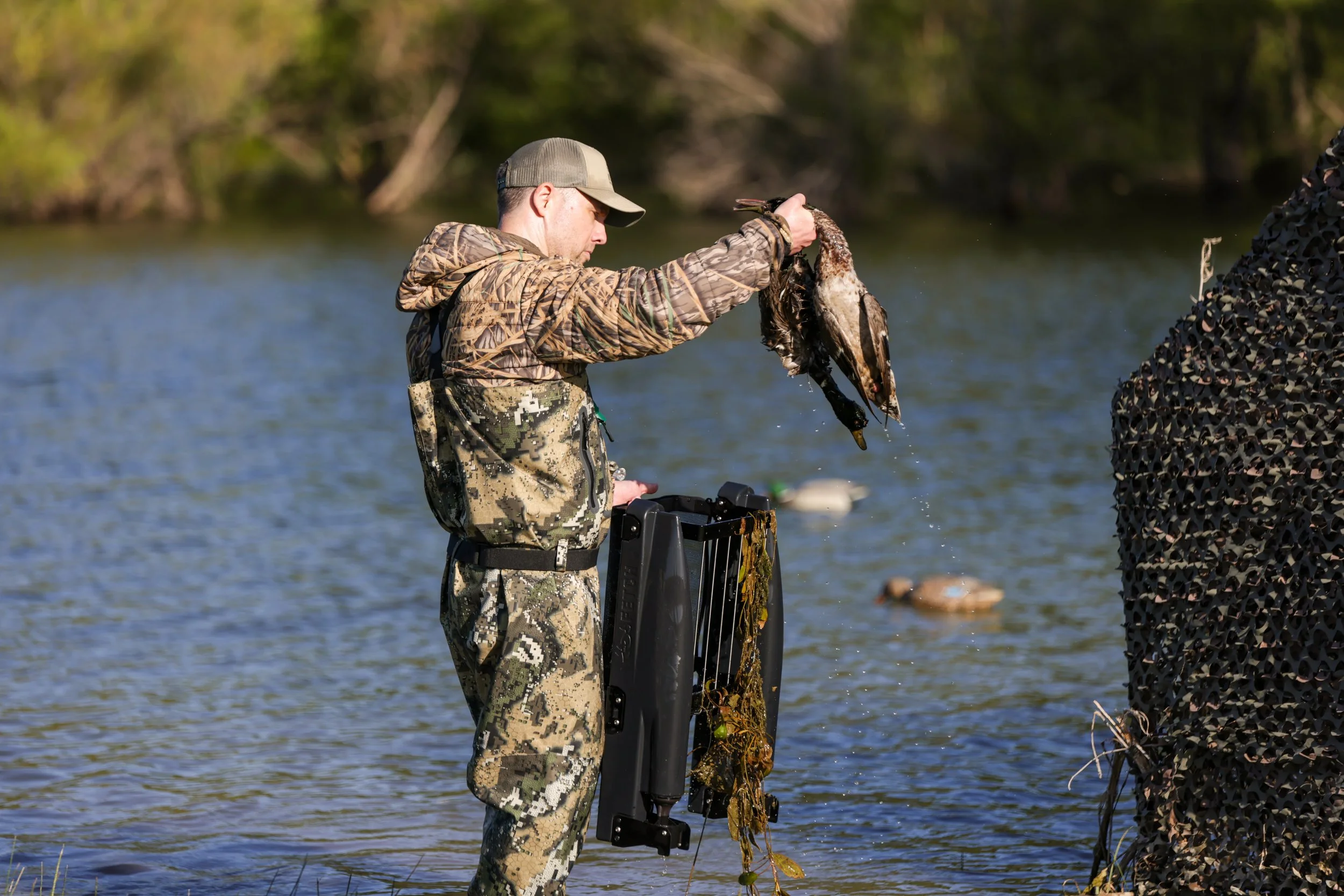 A man in camouflage clothing and a cap is fishing in a body of water, pulling a duck out of the collector of the Fetch Waterfowl Retrieval System
