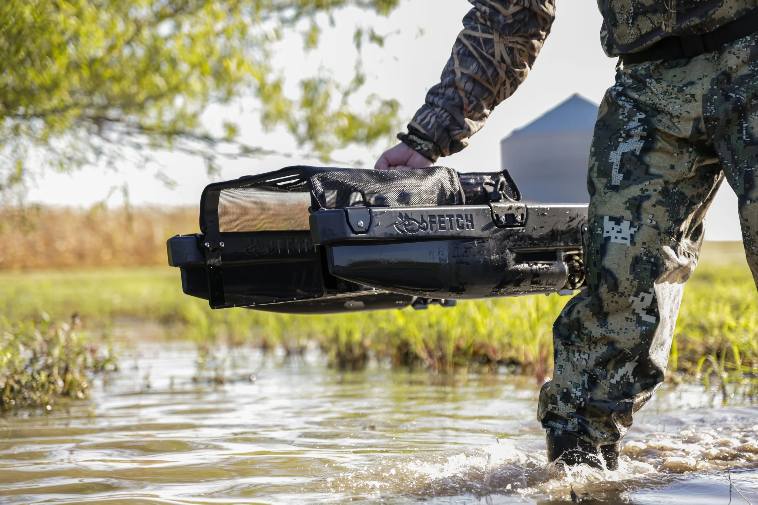 Person wearing camouflage clothing wading through water, holding a Fetch waterfowl retrieval system
