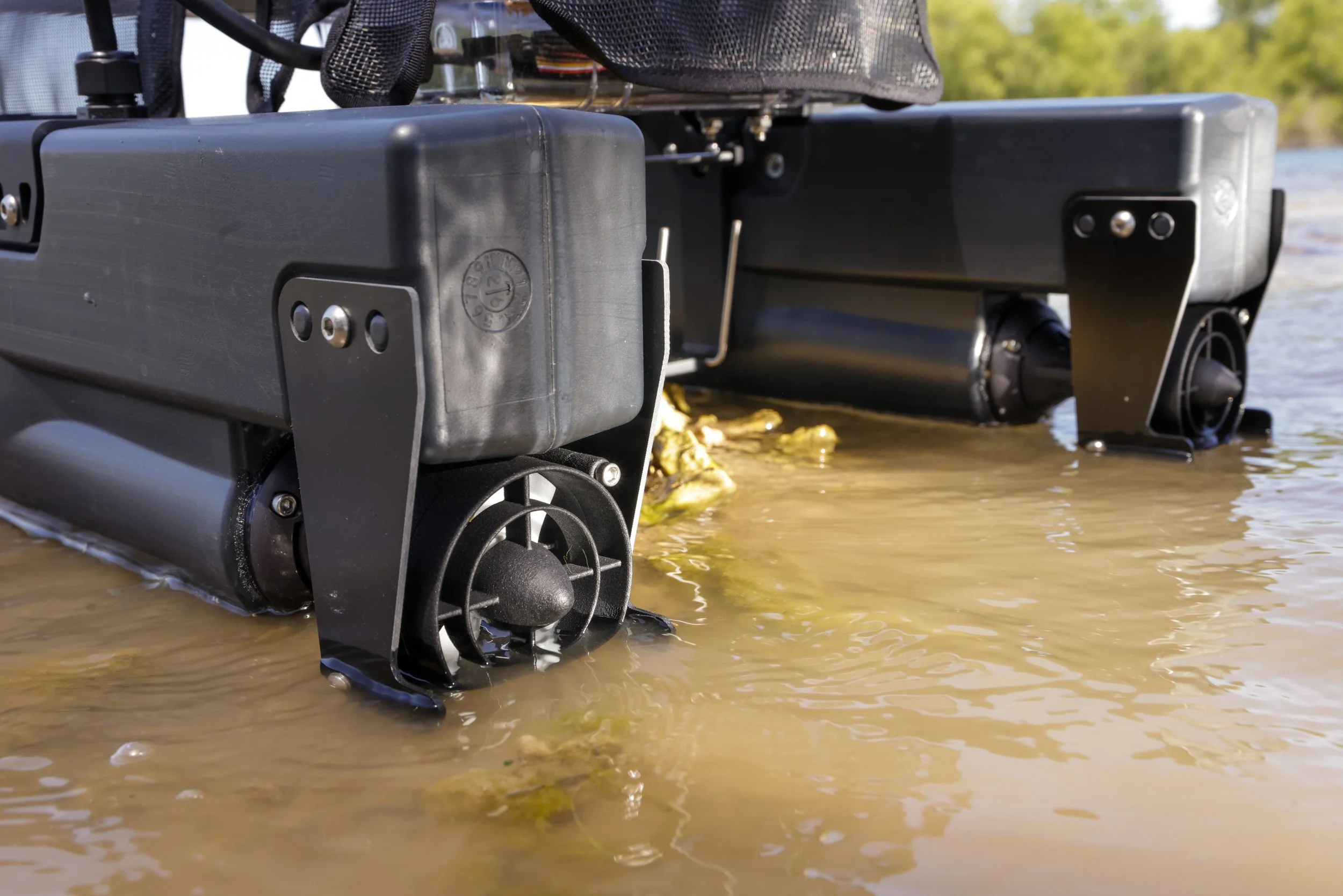 Close-up of a remote-controlled boat with a black hull, partially submerged in a body of water with a sandy shoreline, trees, and a blue sky in the background.