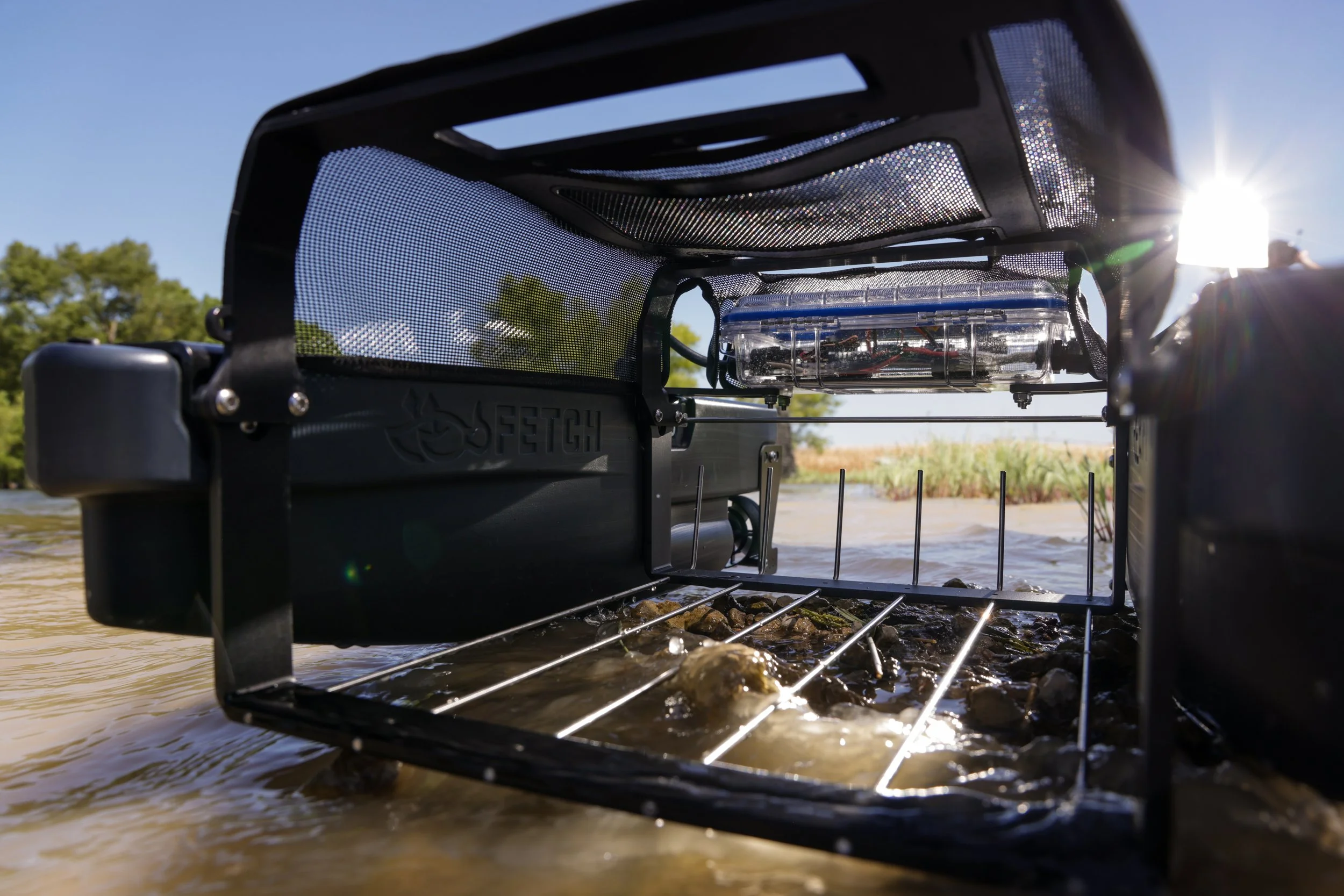 A close-up of the Fetch Waterfowl Retrieval System's waterfowl collection area, situated in a water setting, with the sun shining brightly in the background and a wooded area visible in the distance.