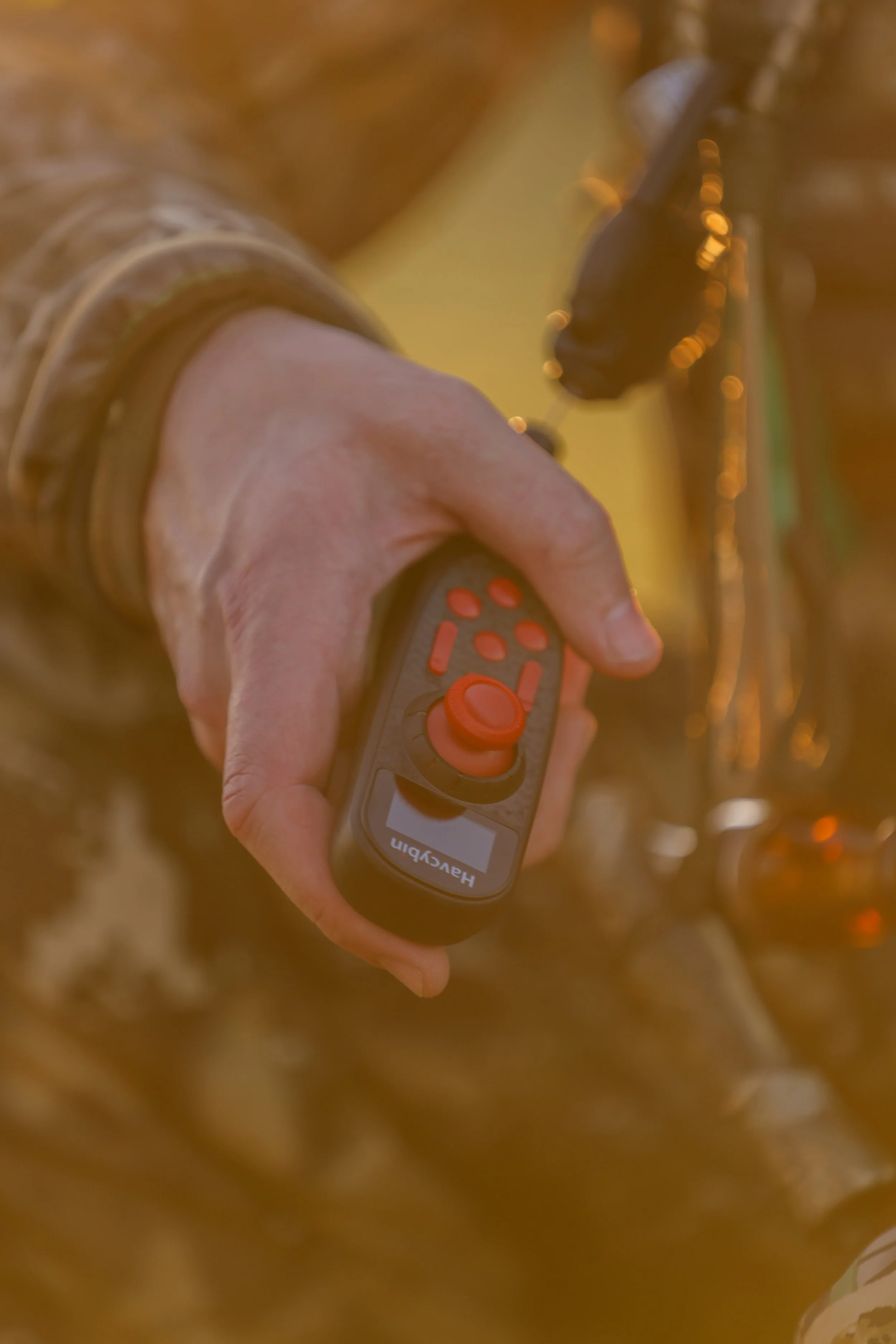 Close-up of a person's hand holding a gray remote control with orange buttons for the Fetch Waterfowl Retrieval System, pointing upwards amidst a blurred outdoor background with warm sunlight.