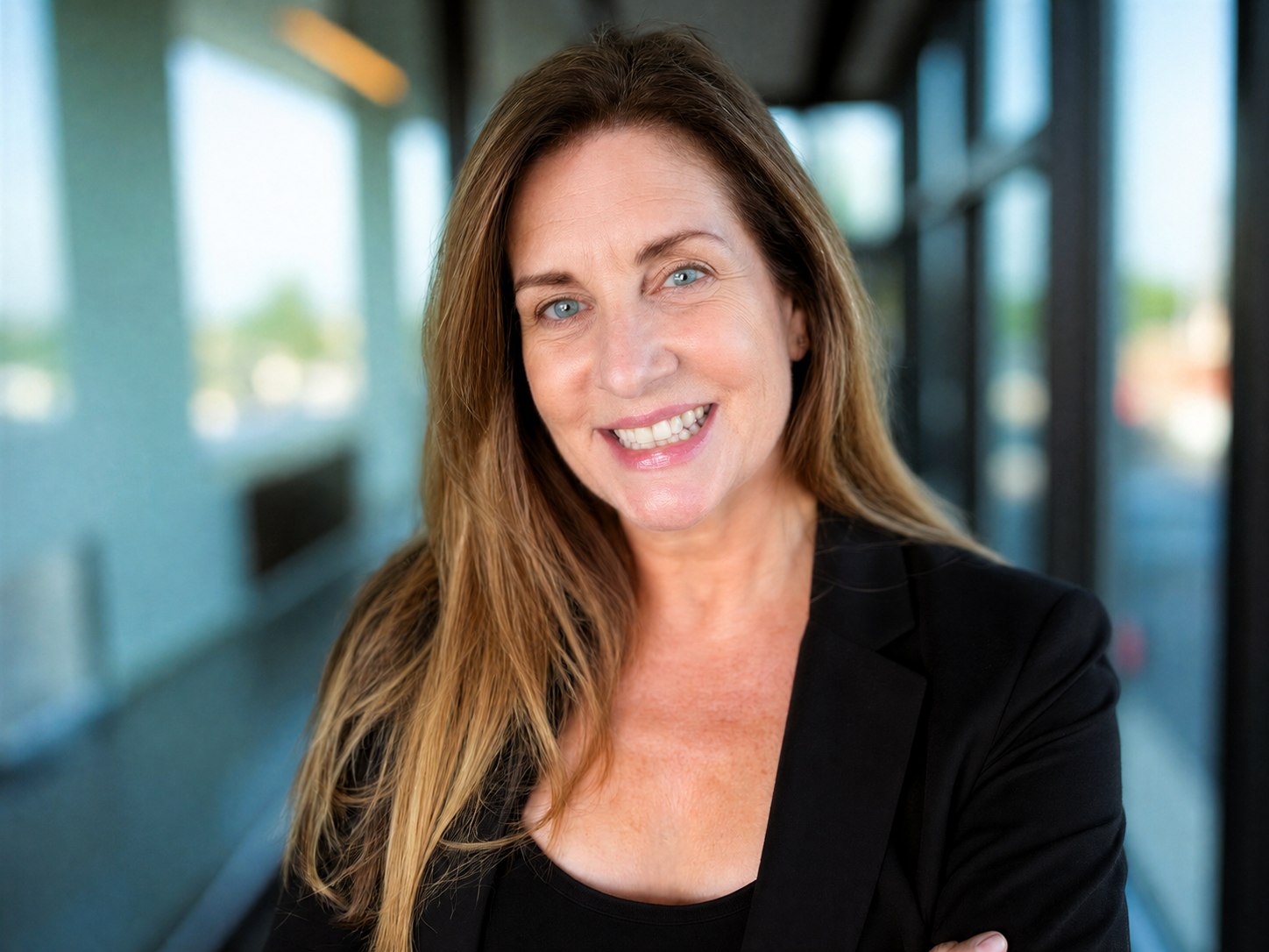A woman with long brown hair and blue eyes smiling at the camera, wearing a black blazer and a black top, standing in a modern building with large windows in the background.