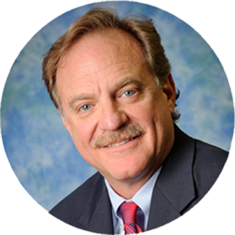 A professional headshot of a middle-aged man with light brown hair, a mustache, and blue eyes, wearing a dark suit, white shirt, and red tie, smiling against a blue and green blurred background.