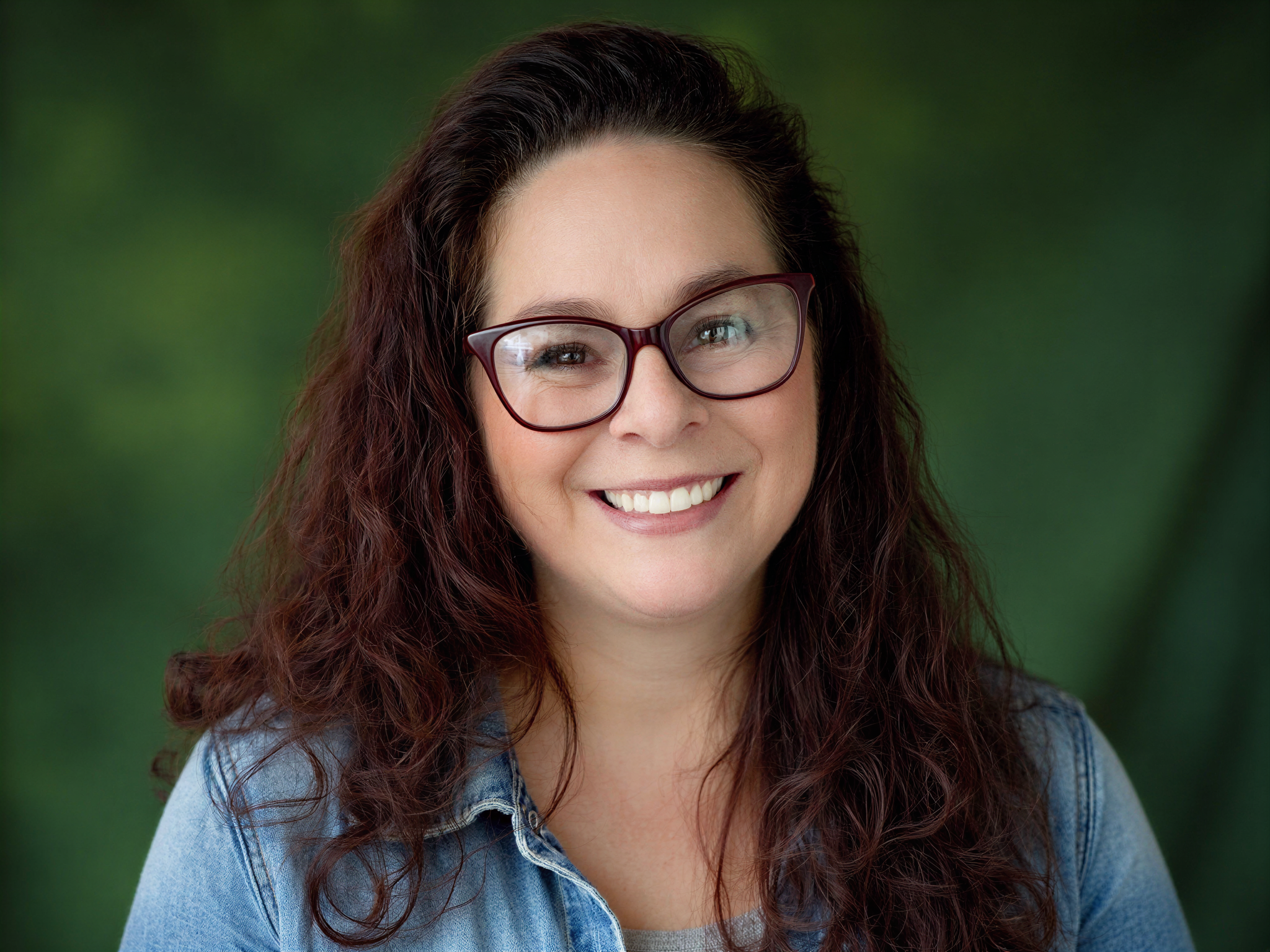 Close-up of a woman with curly reddish-brown hair, wearing glasses and smiling, against a blurred green background.