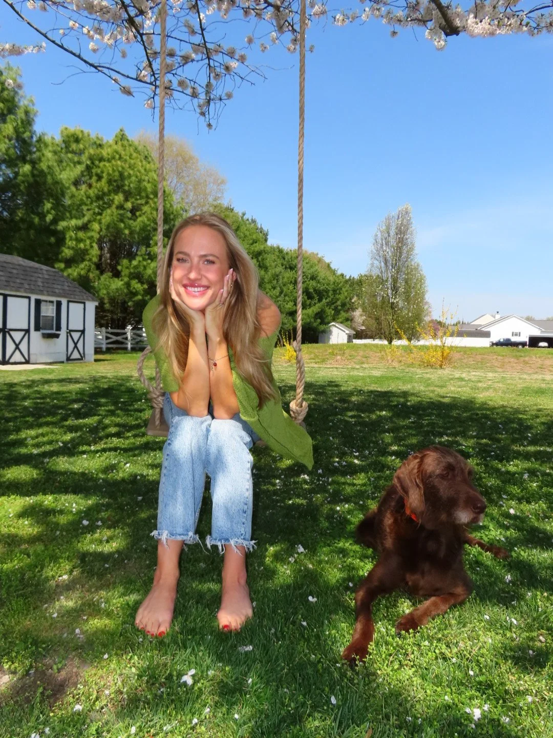 A woman with long blonde hair sitting on a swing hanging from a tree, smiling and resting her face in her hands, while a brown dog runs on the grass nearby on a sunny day with clear blue skies.