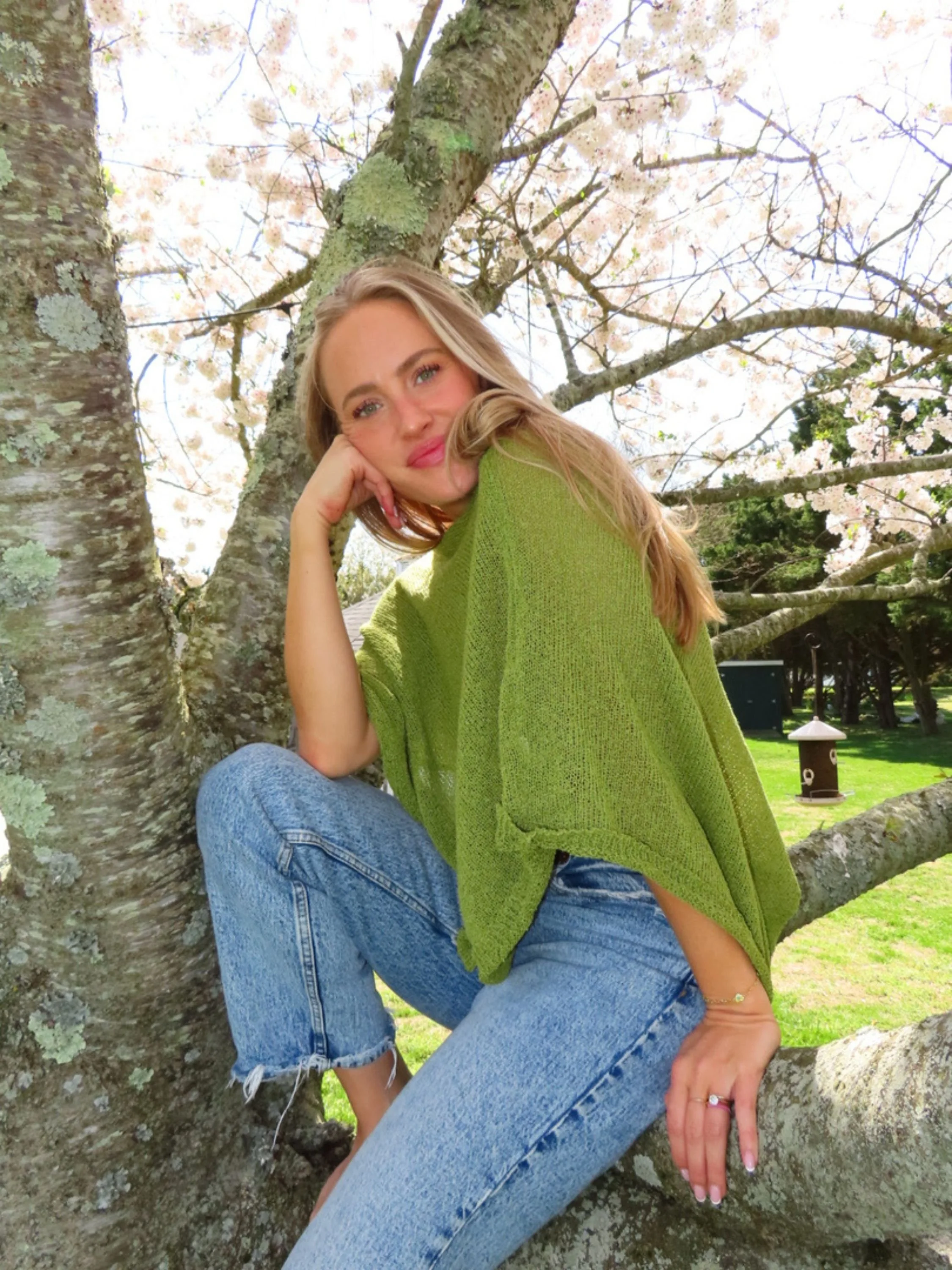 A woman with long blonde hair, wearing a green sweater and blue jeans, sitting on a tree branch outdoors with blooming cherry blossom trees in the background.
