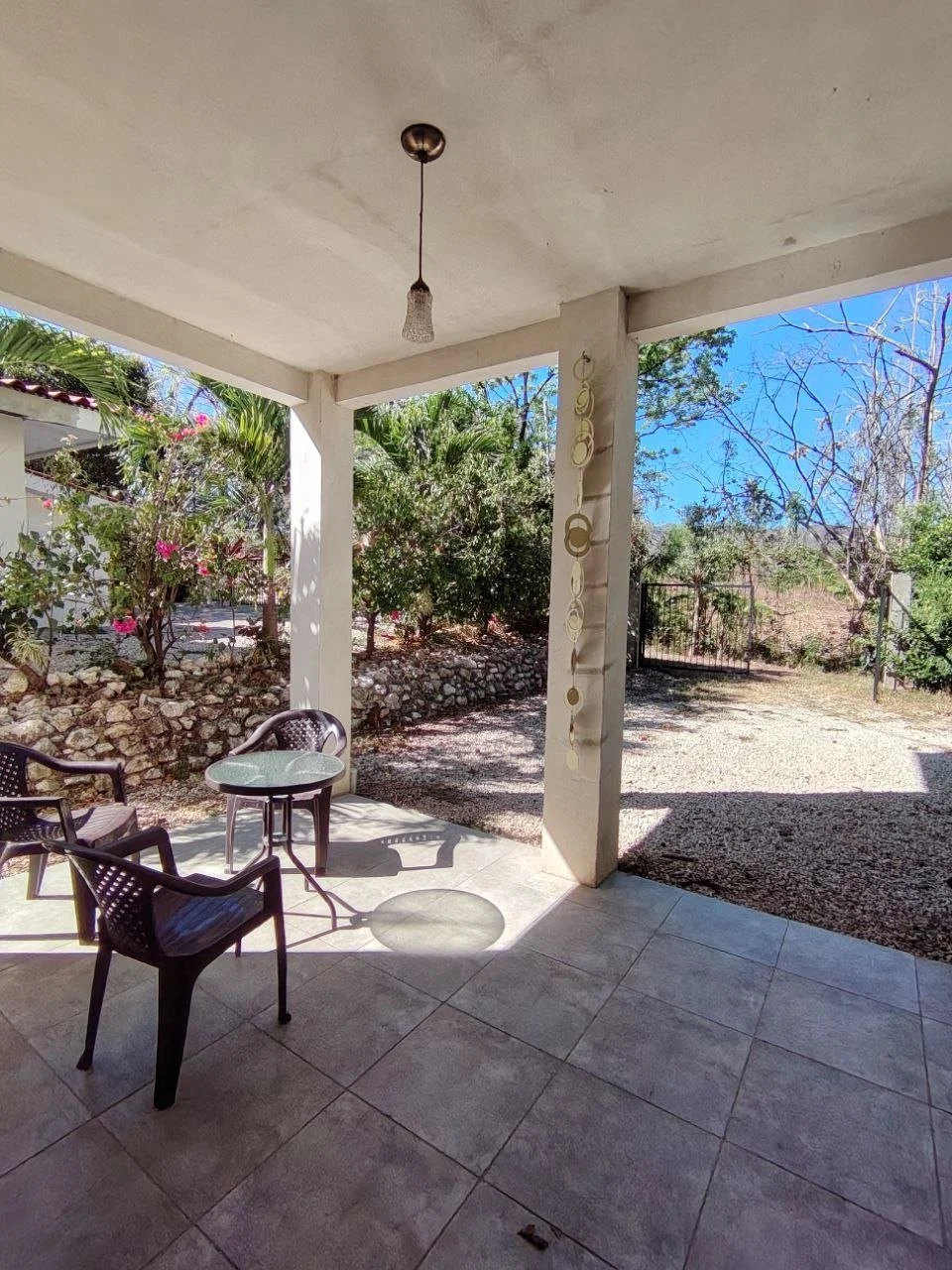 Porch with tiled floor, three black plastic chairs, round glass table, overhead light fixture, view of trees and garden in sunny weather.