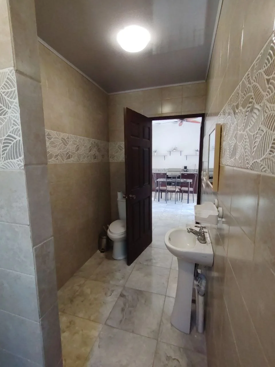 Small bathroom with beige tile walls and floor, a white pedestal sink, a mirror above the sink, a toilet in the corner, and a ceiling light. An open dark wooden door leads to a dining area with a table and chairs visible in the background.