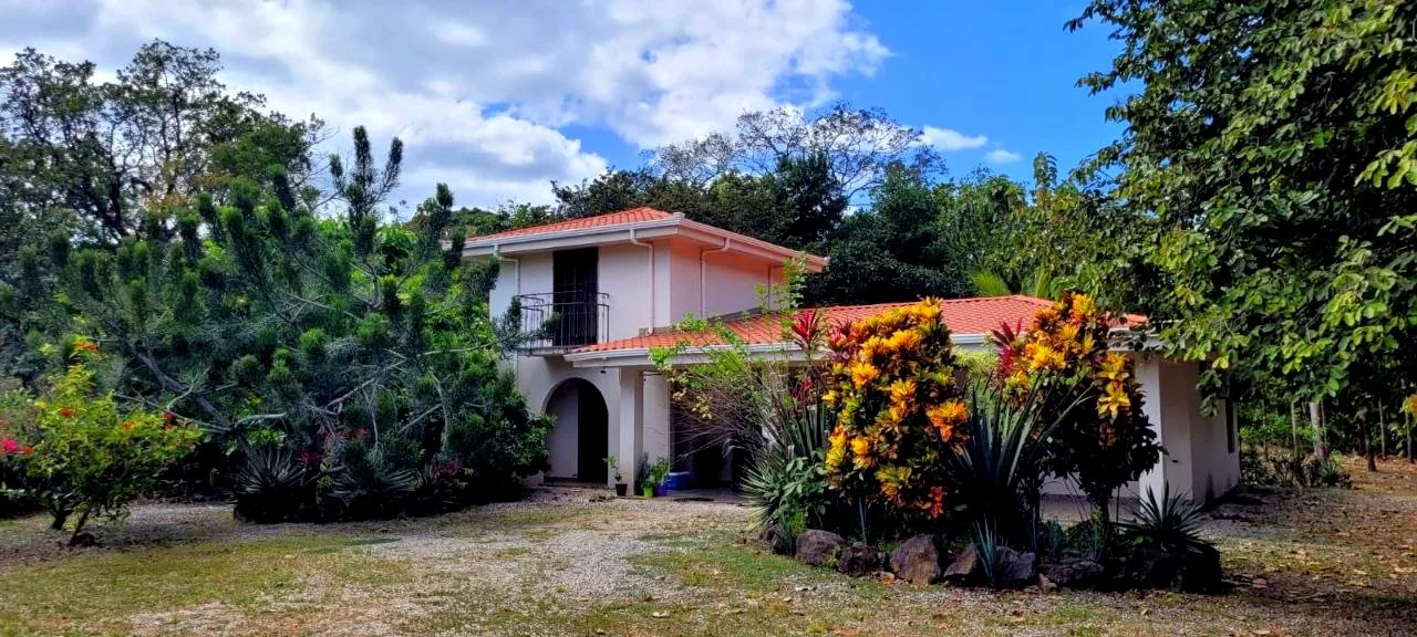 A two-story house with a red tile roof, surrounded by greenery and colorful plants, under a partly cloudy sky.