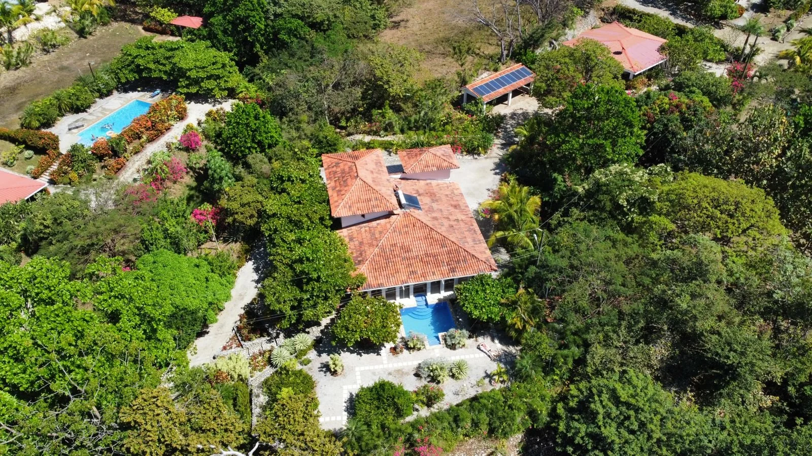 Aerial view of a house with a red-tiled roof, surrounded by lush green trees and shrubbery, with a swimming pool and garden area on the property.