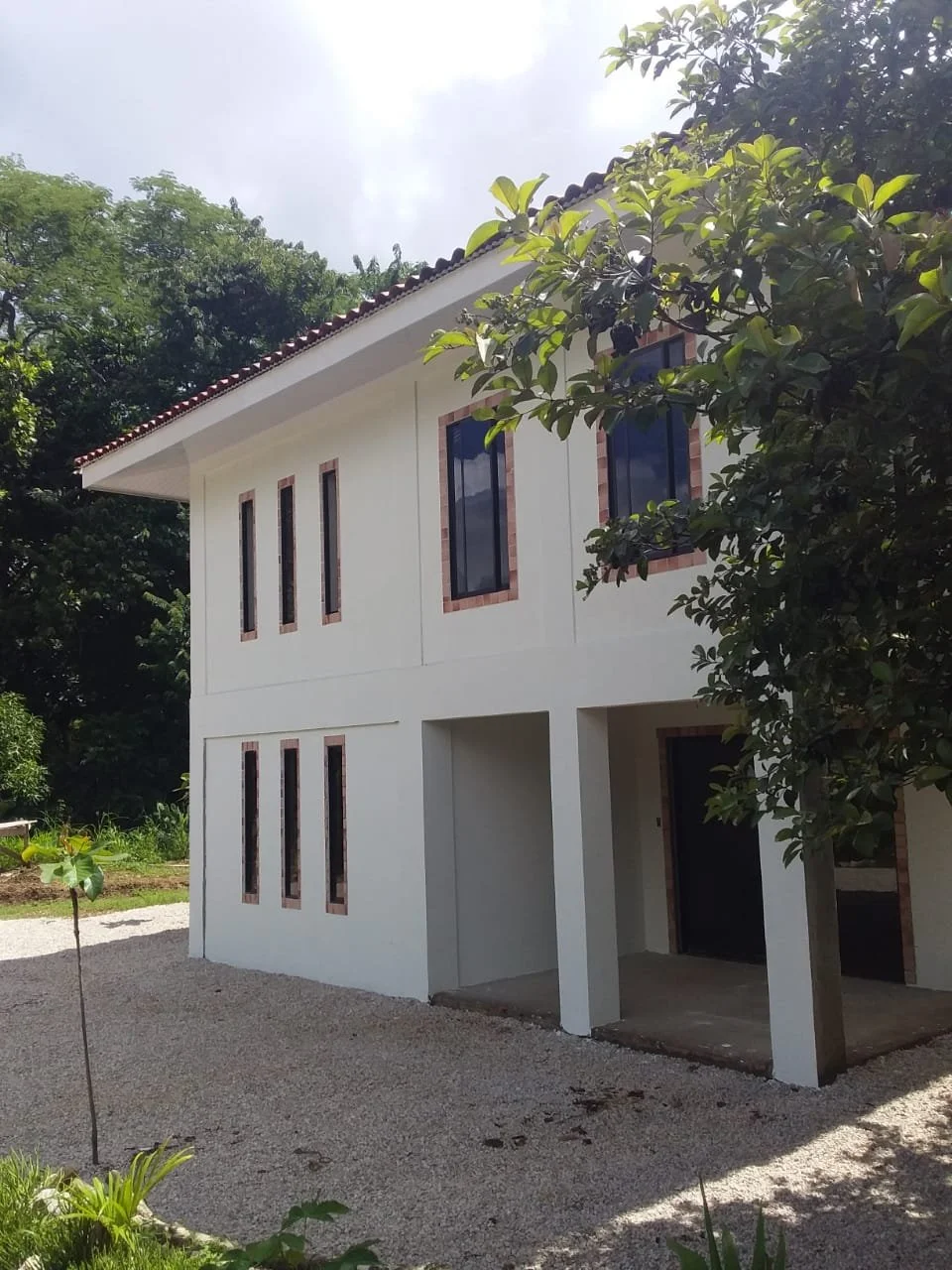 A two-story modern house with white exterior walls, multiple narrow windows with brick borders, and an open garage underneath. Surrounded by trees and greenery.