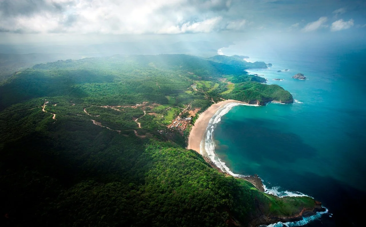 Aerial view of a lush green coastal landscape with a sandy beach and rocky outcroppings along the ocean, under a cloudy sky.