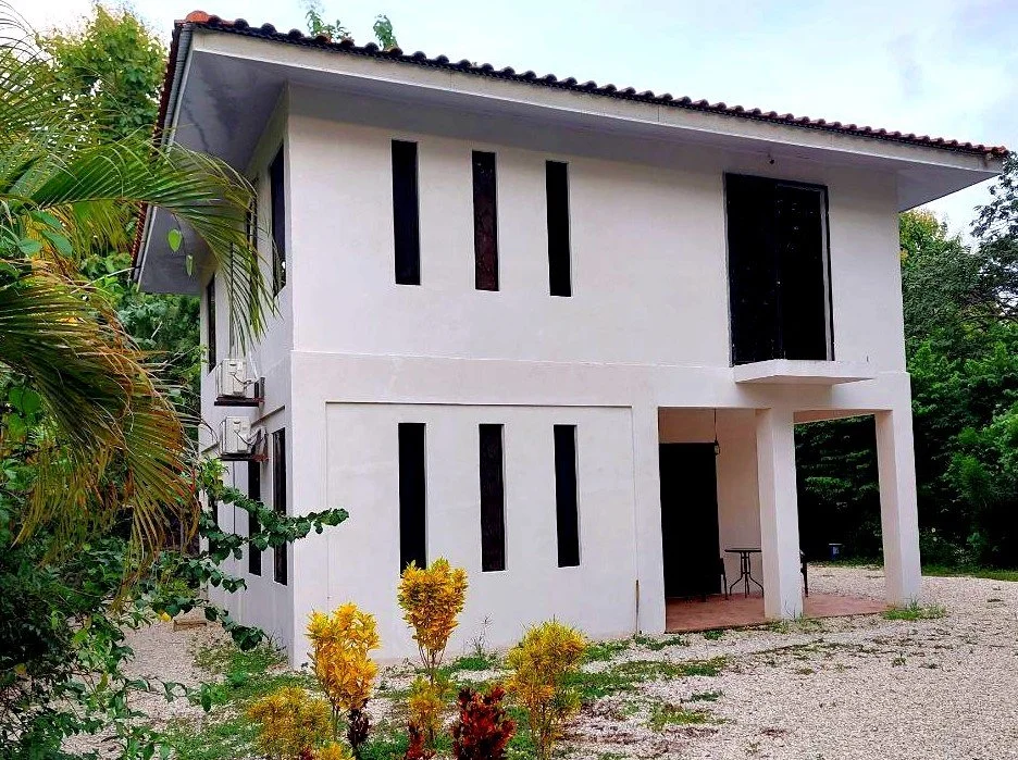 Modern two-story white house with a sloped roof and small window openings, surrounded by greenery and a gravel yard.