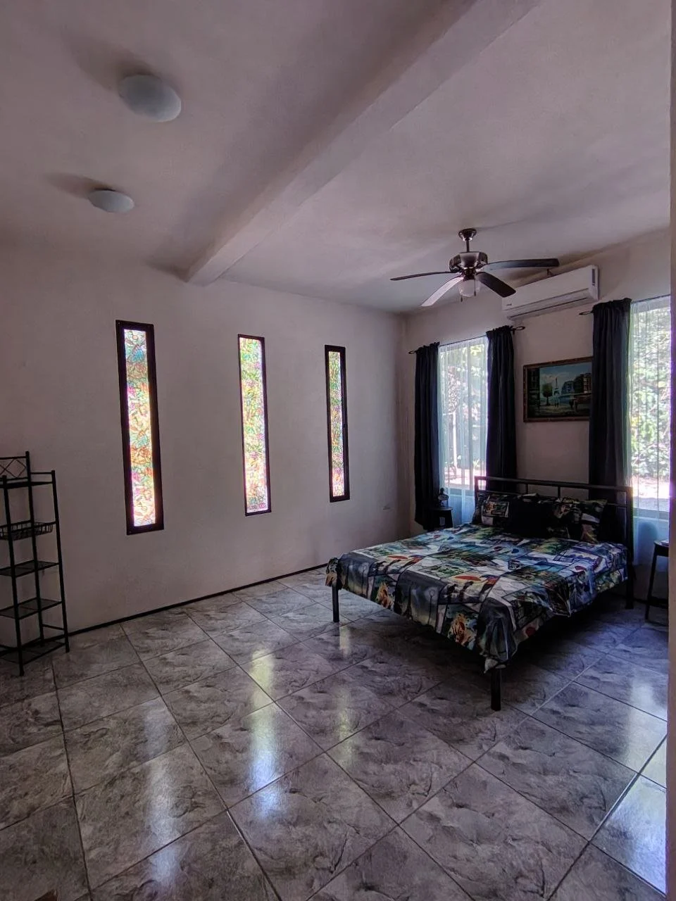 A bedroom with a bed covered in a colorful patterned comforter, dark curtains on windows, a ceiling fan, and tiled flooring.