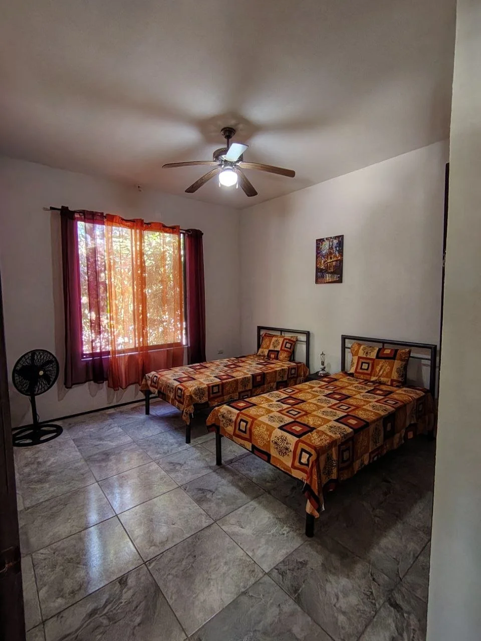 A bedroom with two twin beds covered with patterned orange, brown, and beige bedspreads, a window with purple and orange curtains letting in natural light, a standing fan, a ceiling fan with lights, and a small painting on the wall.