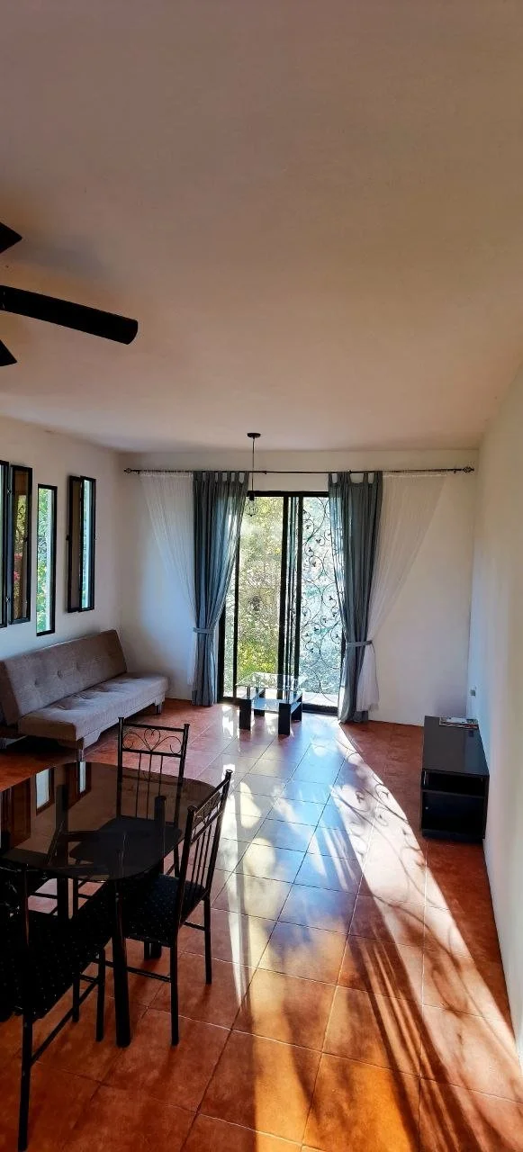 Living room with a beige sofa, black metal dining table with four chairs, tile floors, large windows with curtains, and sunlight casting shadows.