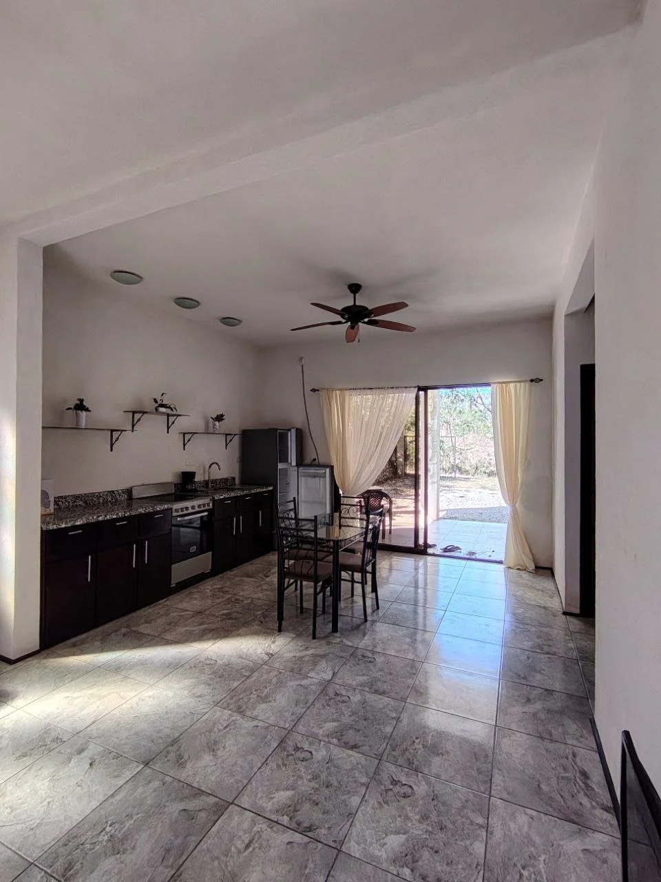 Empty kitchen and dining area with tiled floor, dark cabinets, a round table with chairs, and a sliding glass door with curtains leading outside.