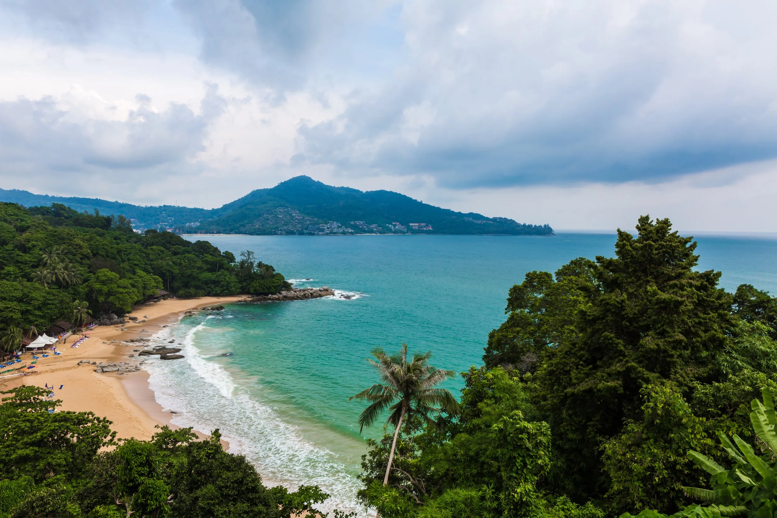 A tropical beach with palm trees, white sand, and turquoise water, backed by lush green hills and a mountain under a partly cloudy sky.
