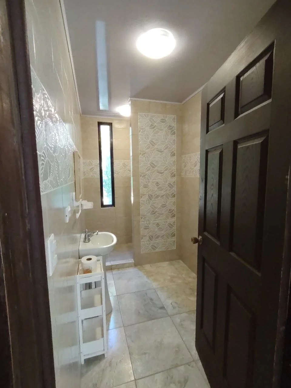 Bathroom with beige tiled walls and floor, small sink, narrow vertical window, decorative tile panel, open dark wooden door.
