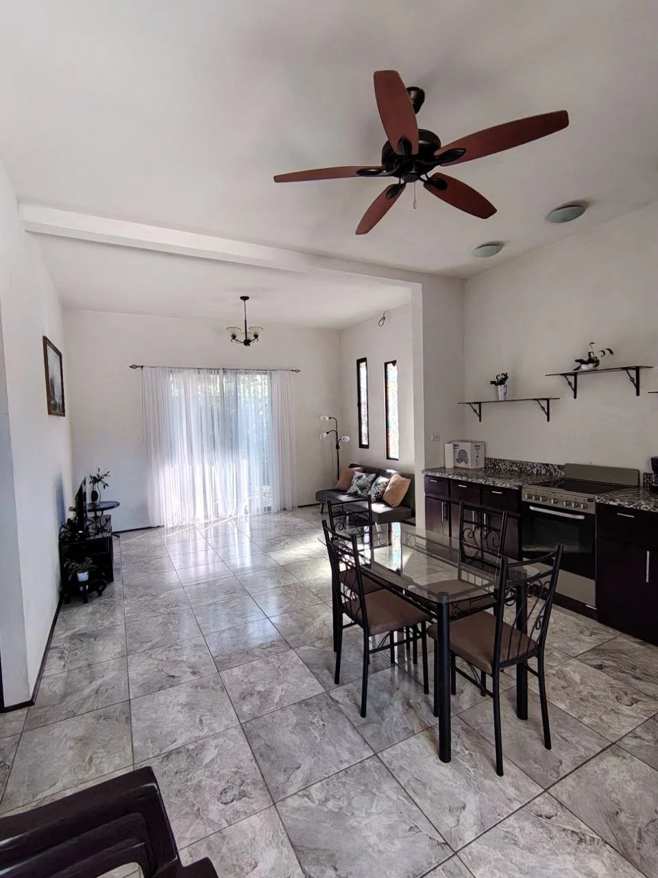 Open-concept living room and kitchen with tiled floor, large window with sheer curtains, ceiling fan, chandelier, small sofa, dining table with four chairs, and kitchen cabinets with open shelves.
