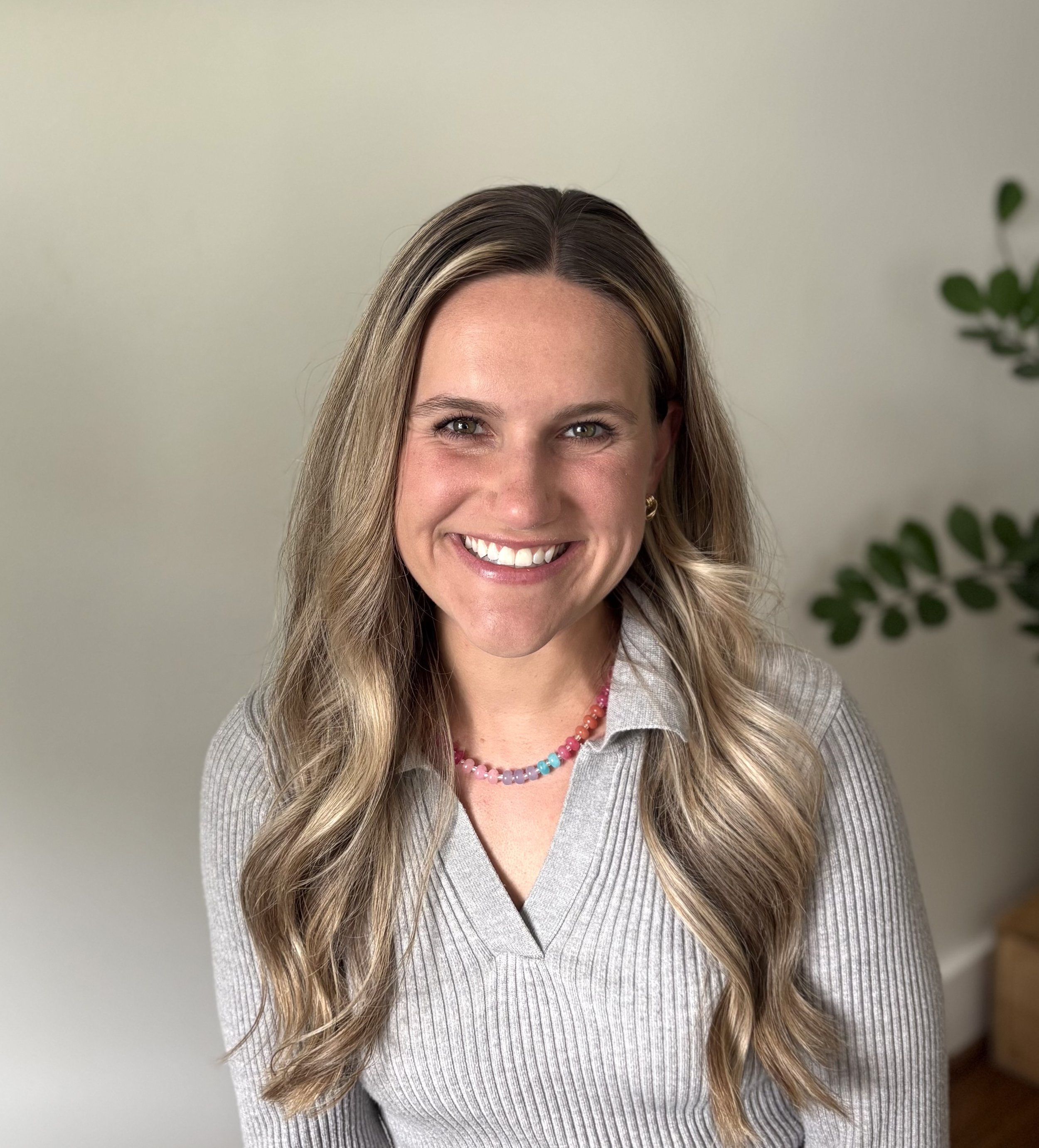 Close-up of a smiling woman with long, wavy blonde hair, wearing a striped gray top and a multicolored beaded necklace, standing in front of a plain light-colored wall and a partially visible green plant.