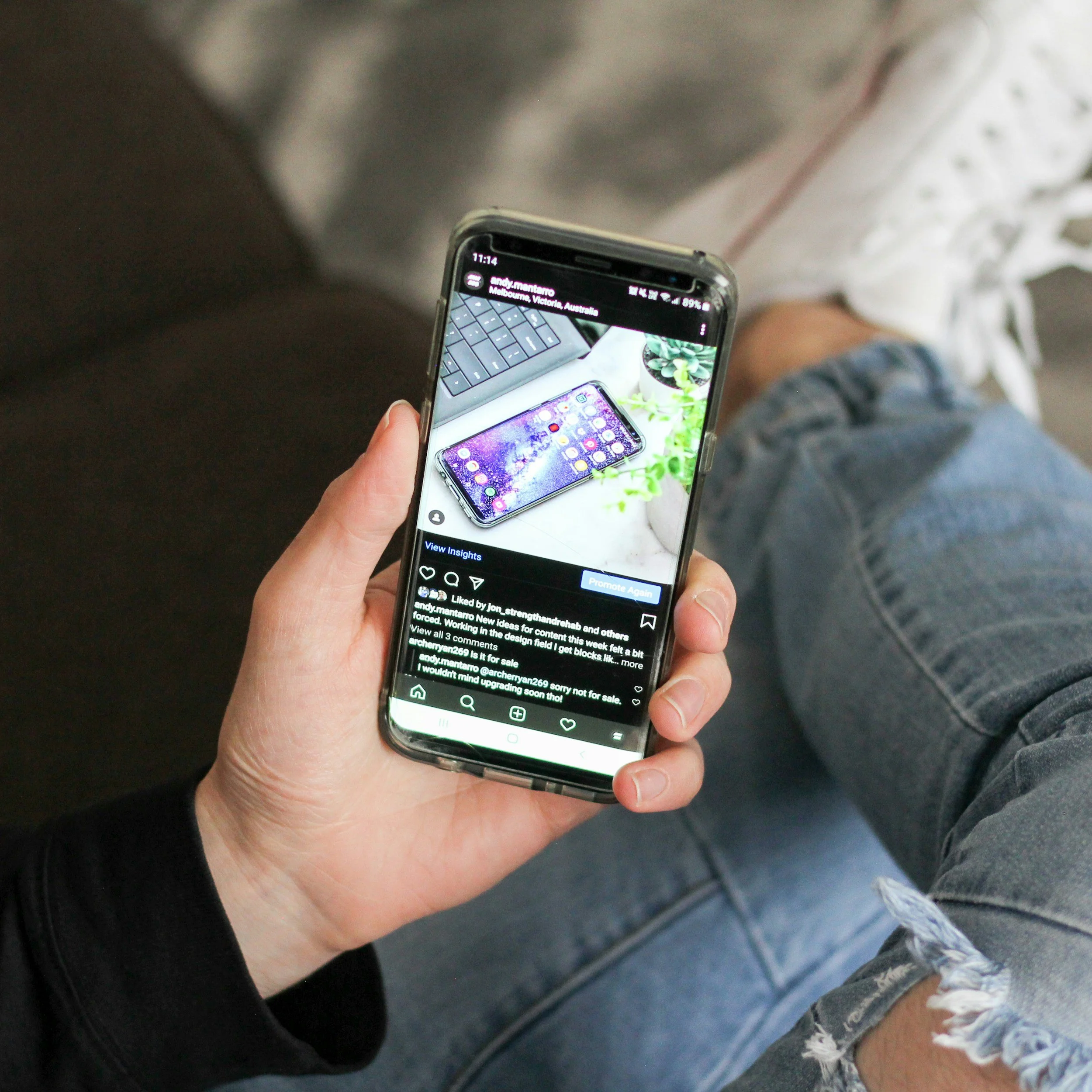 A person holding a smartphone with a social media post visible on the screen. The post shows a decorated phone, possibly in anticipation of a new device release, with a background of a keyboard and plant. The person is wearing jeans and is sitting, with part of a black jacket and their hand visible.
