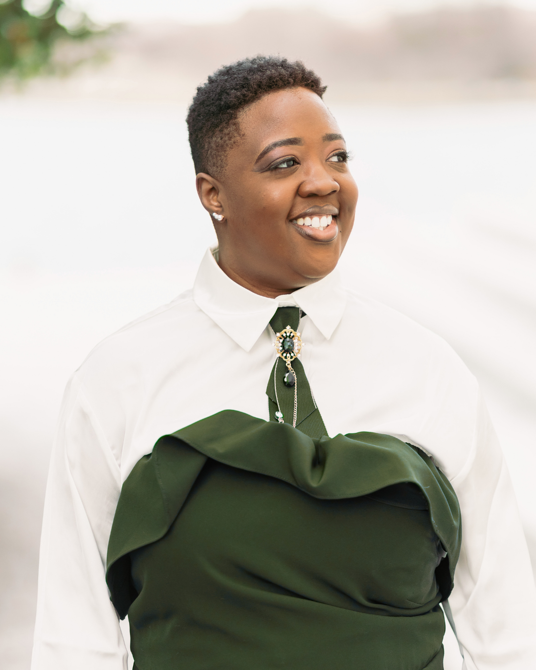 A smiling African American woman (Darrah Ferguson) with short hair dressed in a white blouse and a dark green wrap, standing outdoors near a body of water with blurred trees in the background.