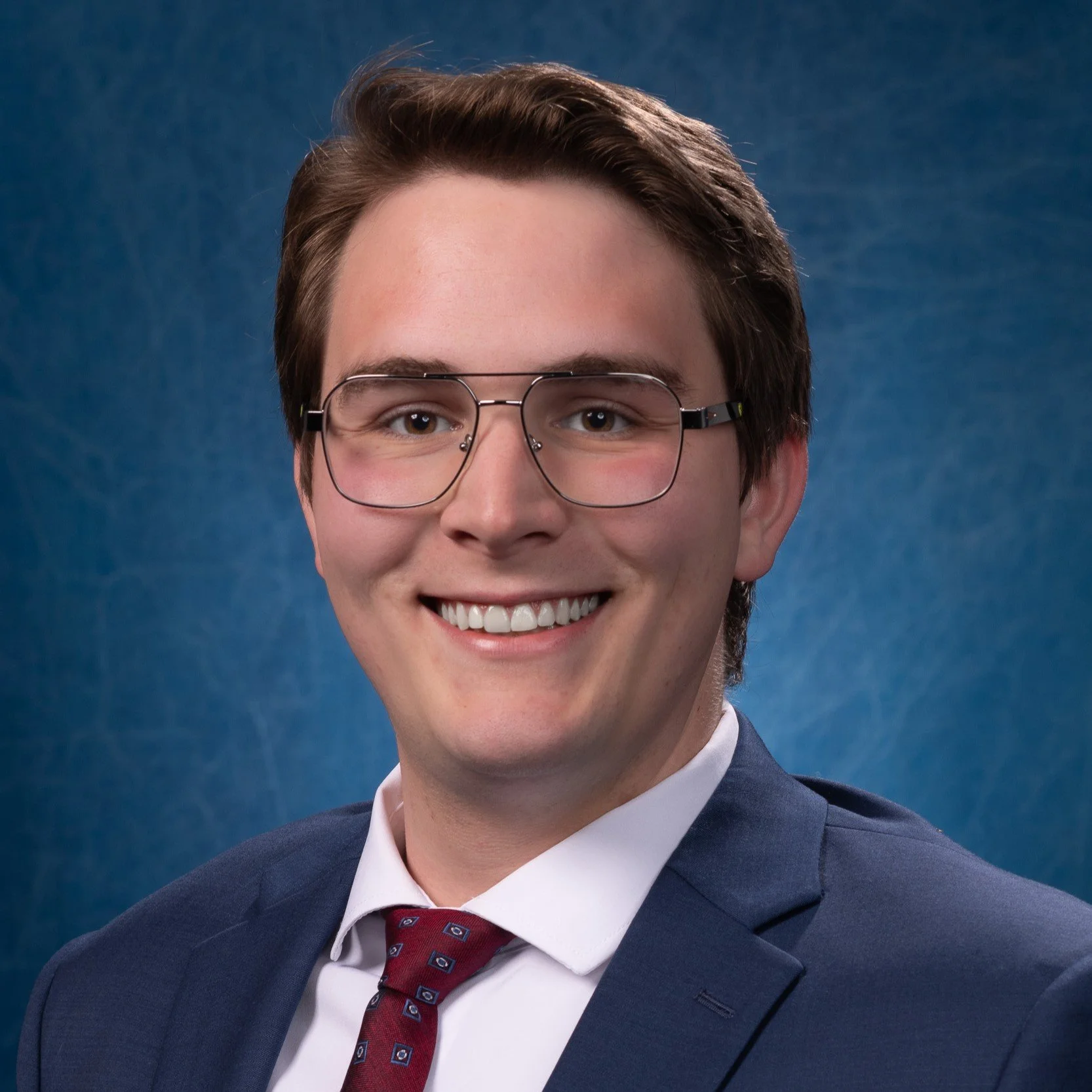 A young man with brown hair and glasses, wearing a suit and tie, smiling against a blue background.