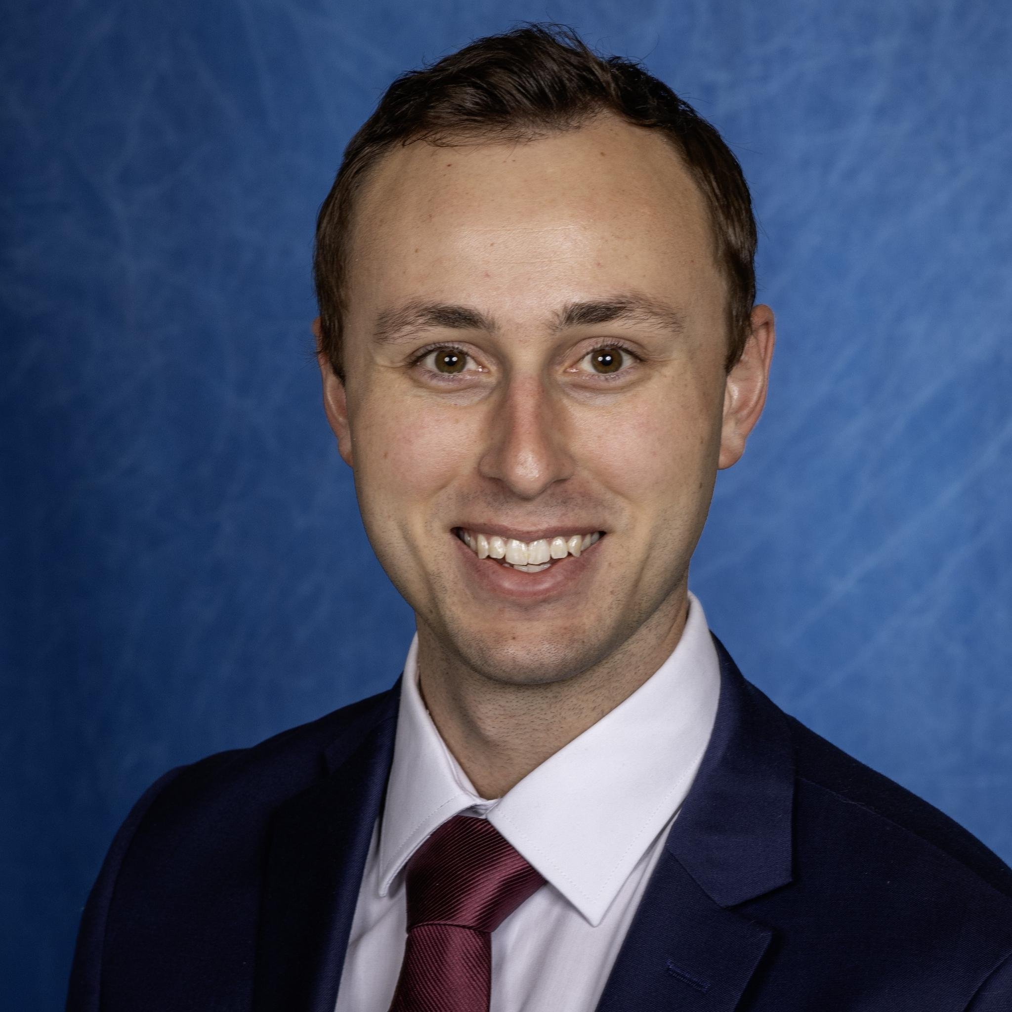 Close-up of a smiling man with short light brown hair, wearing a navy suit, white shirt, and maroon tie, against a blue background.