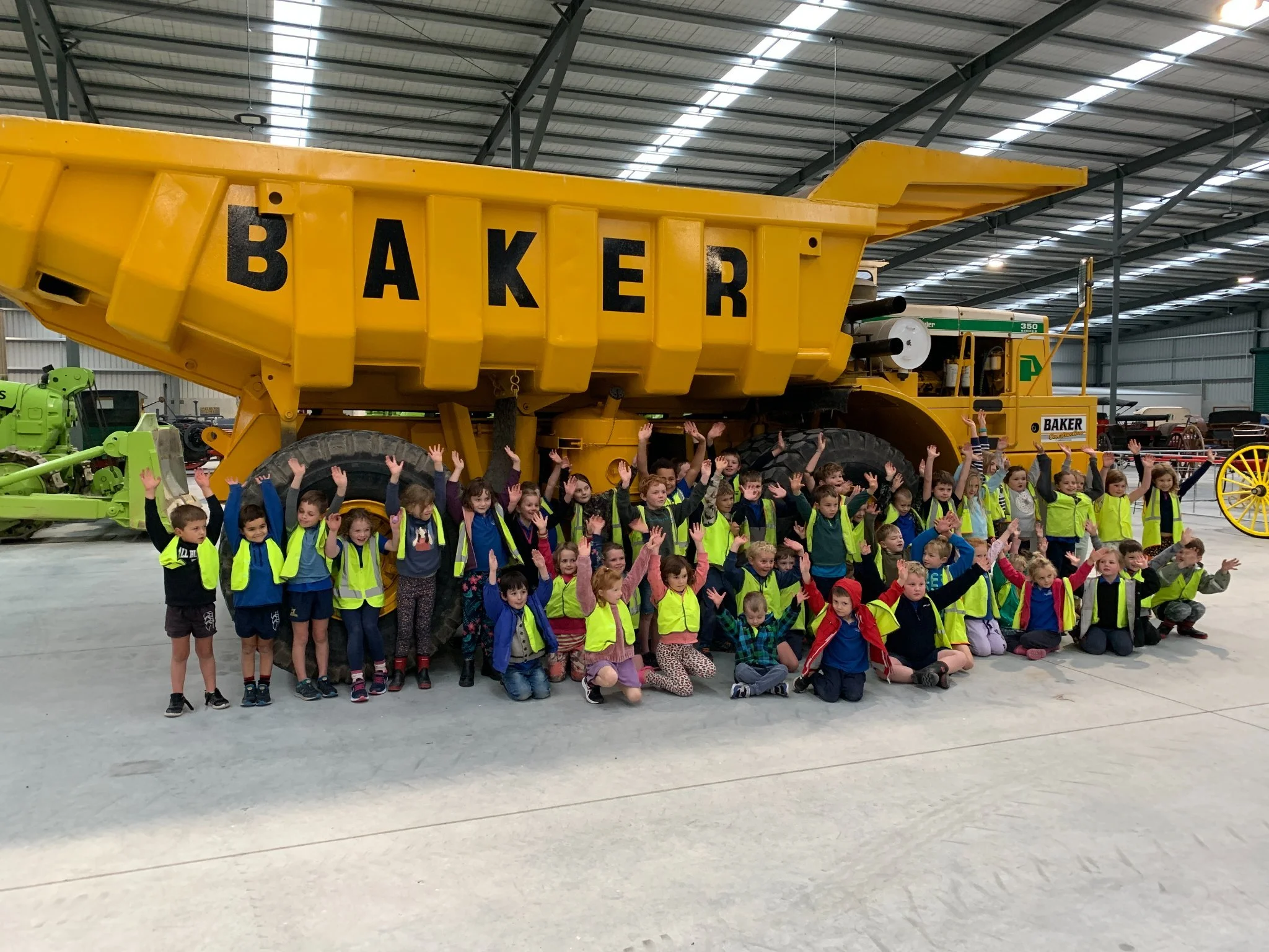 A large group of children, some wearing bright yellow safety vests, gathered inside a spacious warehouse, standing in front of a giant yellow construction truck with the word 'BAKER' painted on it, with their hands raised.