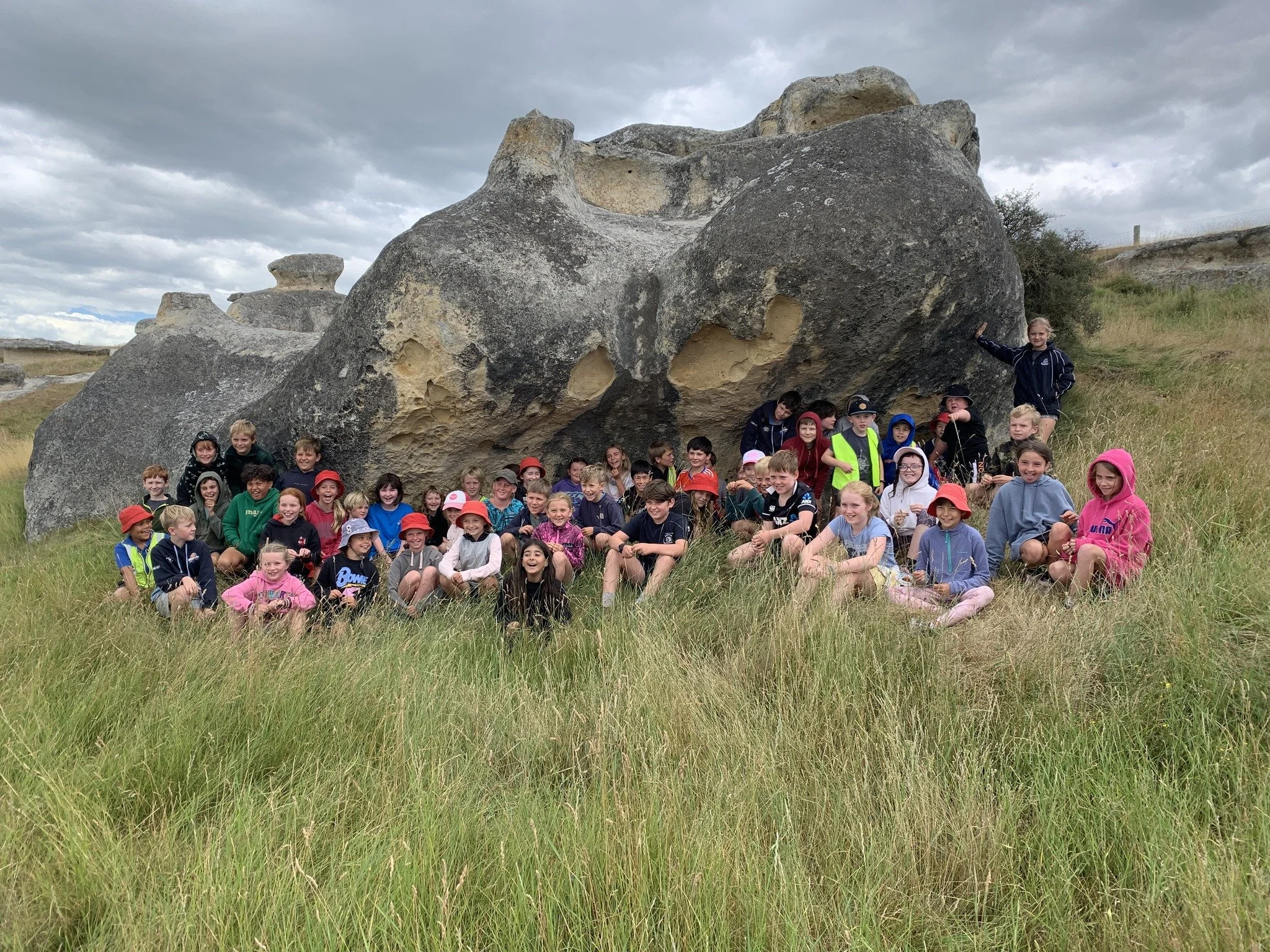 A group of children and a few adults sitting and standing in front of large rock formations outdoors on a cloudy day.