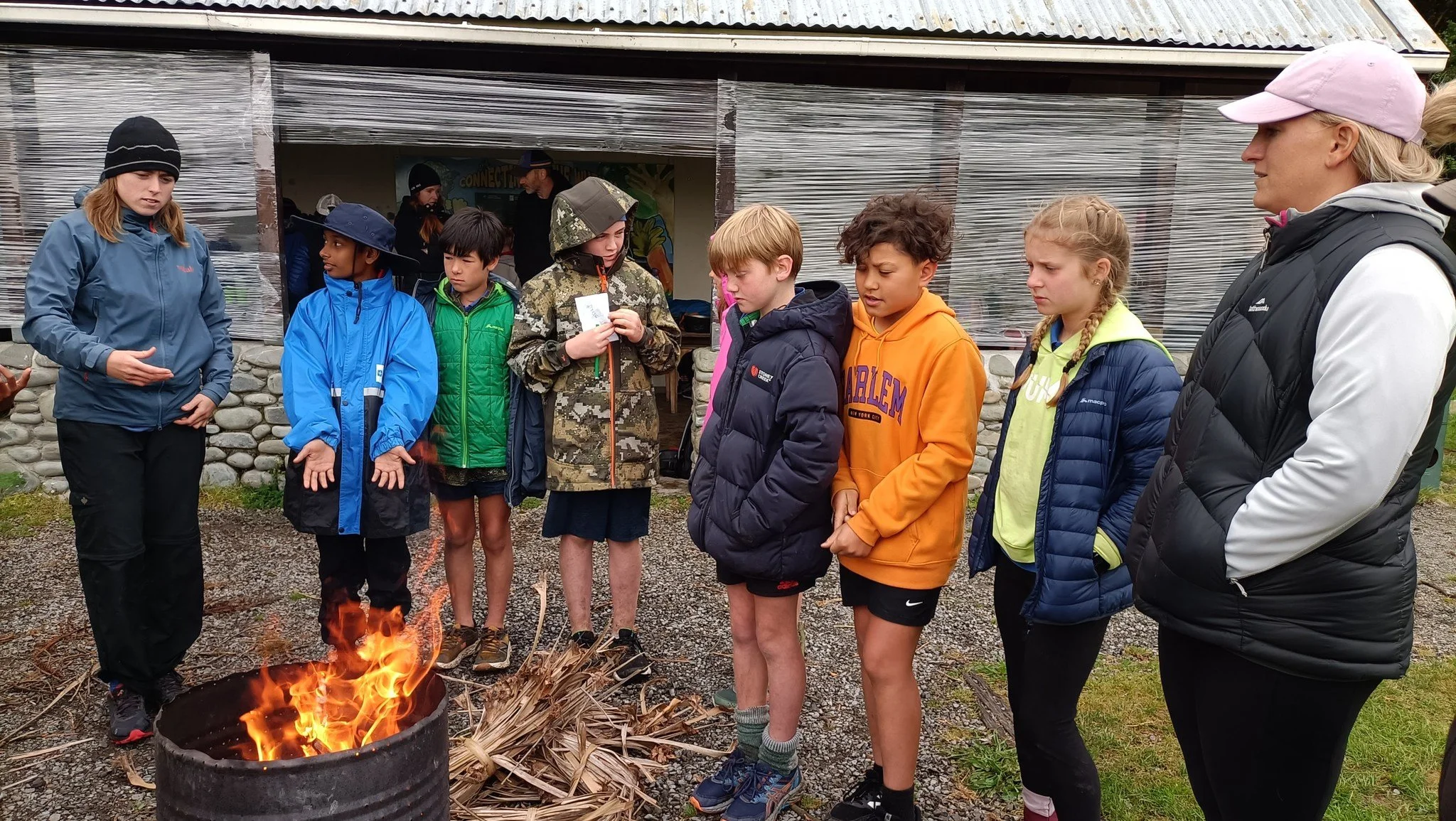 A group of children and two adults standing around a small fire outdoors, with a building and a person in the background.