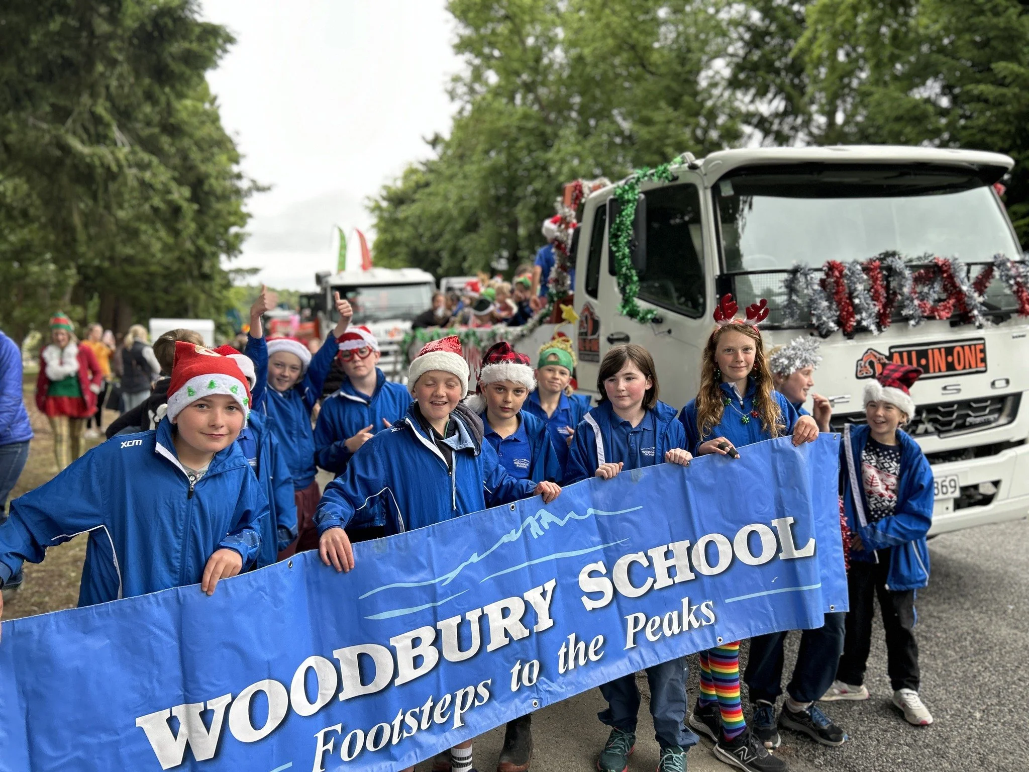 Children wearing Santa hats and reindeer antlers participate in a parade, holding a blue banner that reads "FOODSTEPS to the FOOTHILLS." Behind them, a float decorated with red, green, and silver tinsel is visible, with people sitting on it and a whi