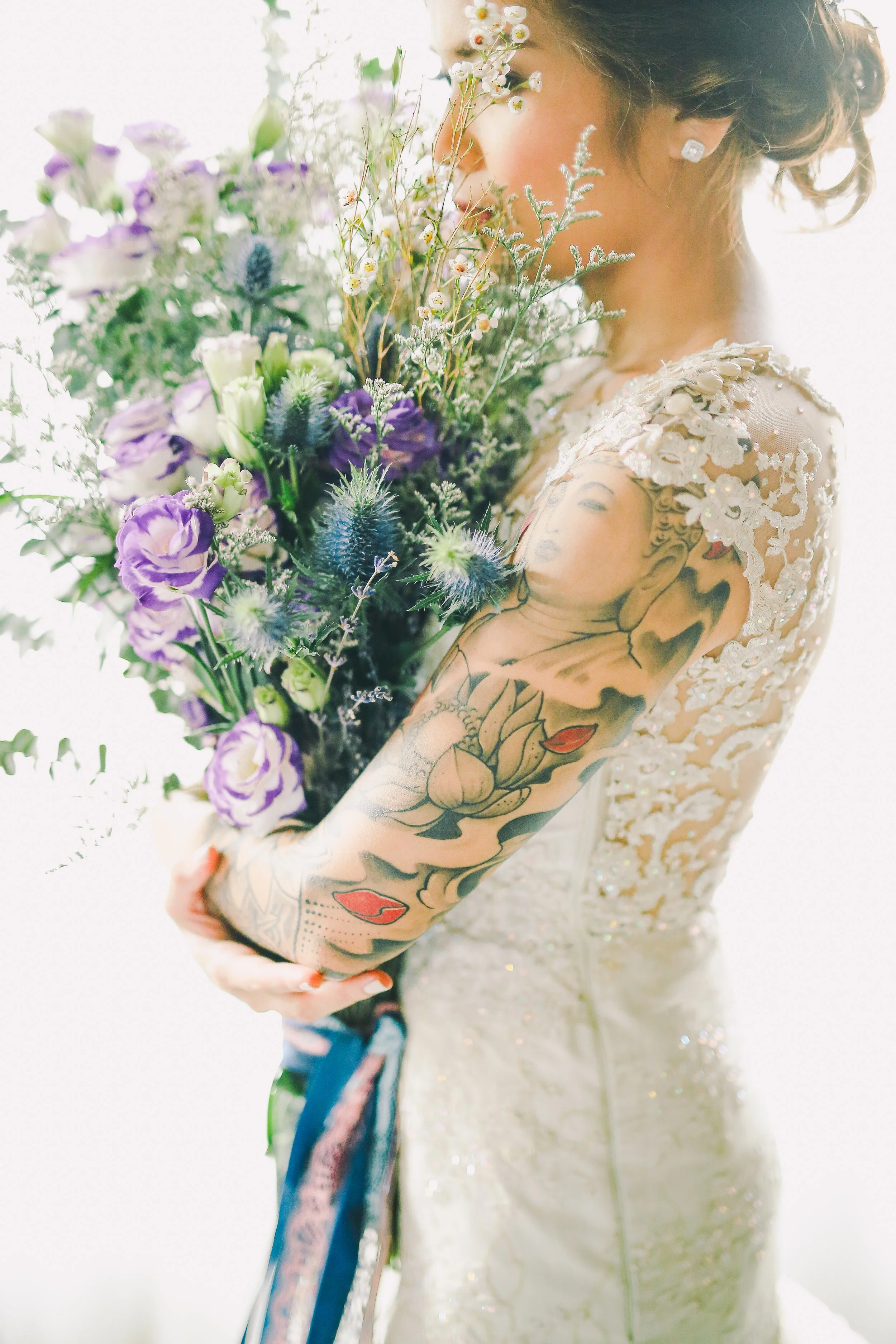 A woman in a wedding dress holding a large bouquet of purple, white, and green flowers with tattoos on her arm. The dress has floral lace and beadwork.