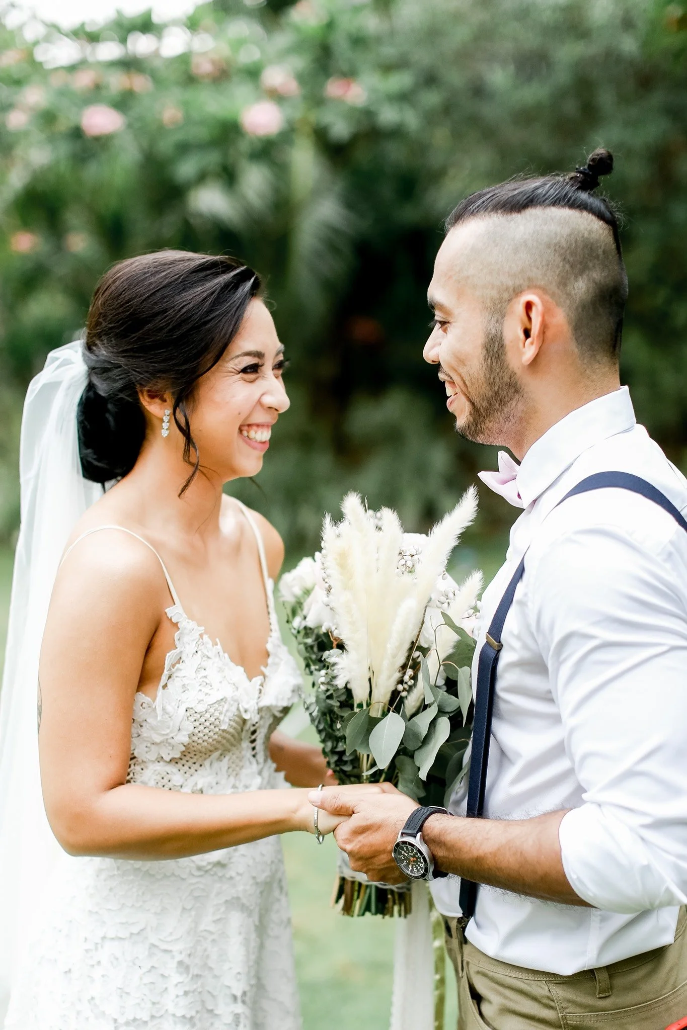 A bride and groom face each other, smiling at their wedding ceremony outdoors, holding hands, with a bouquet of white flowers and greenery.