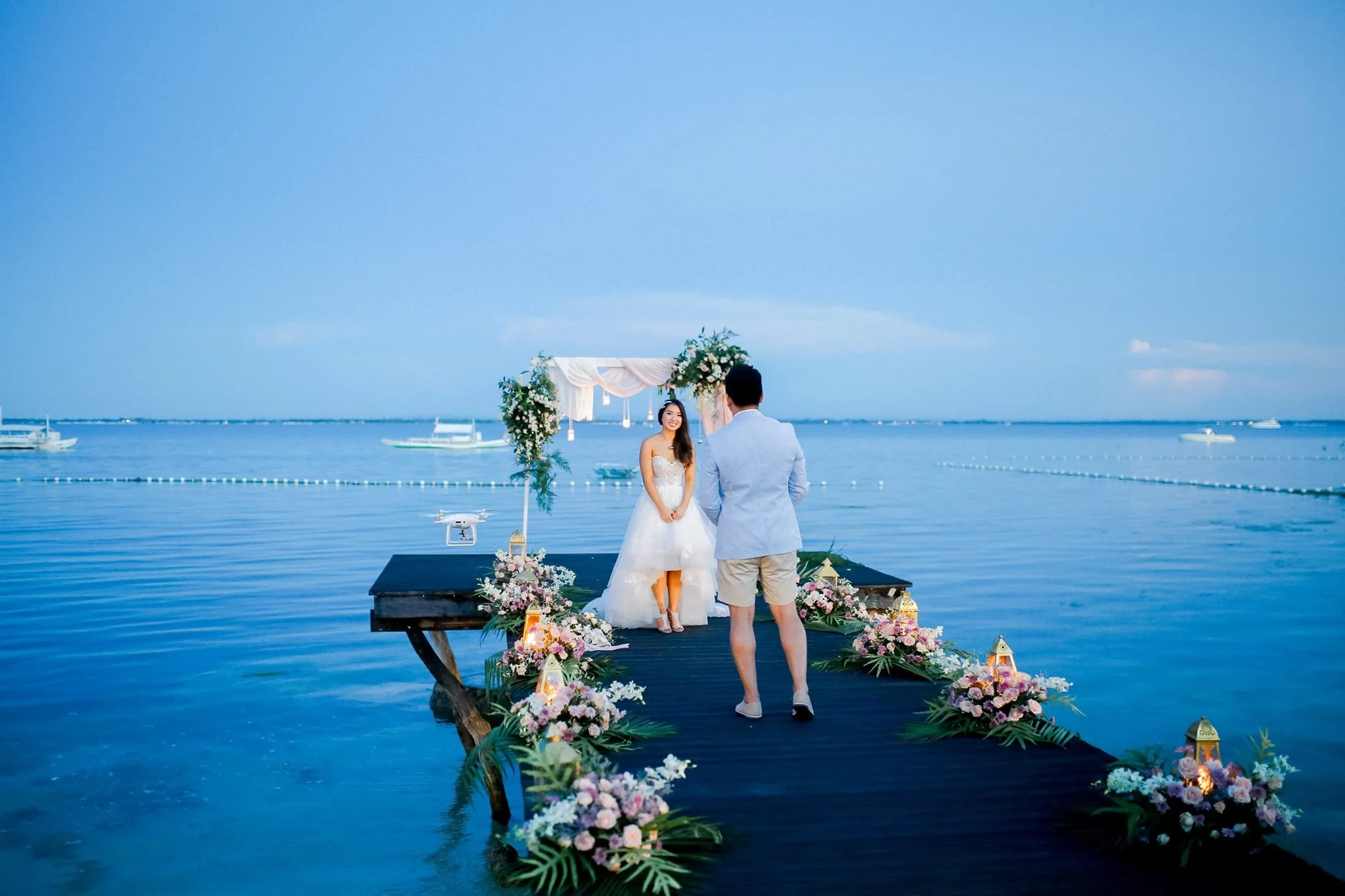 A couple getting married on a wooden dock over calm water with boats in the distance, decorated with floral arrangements and lanterns, against a clear blue sky.