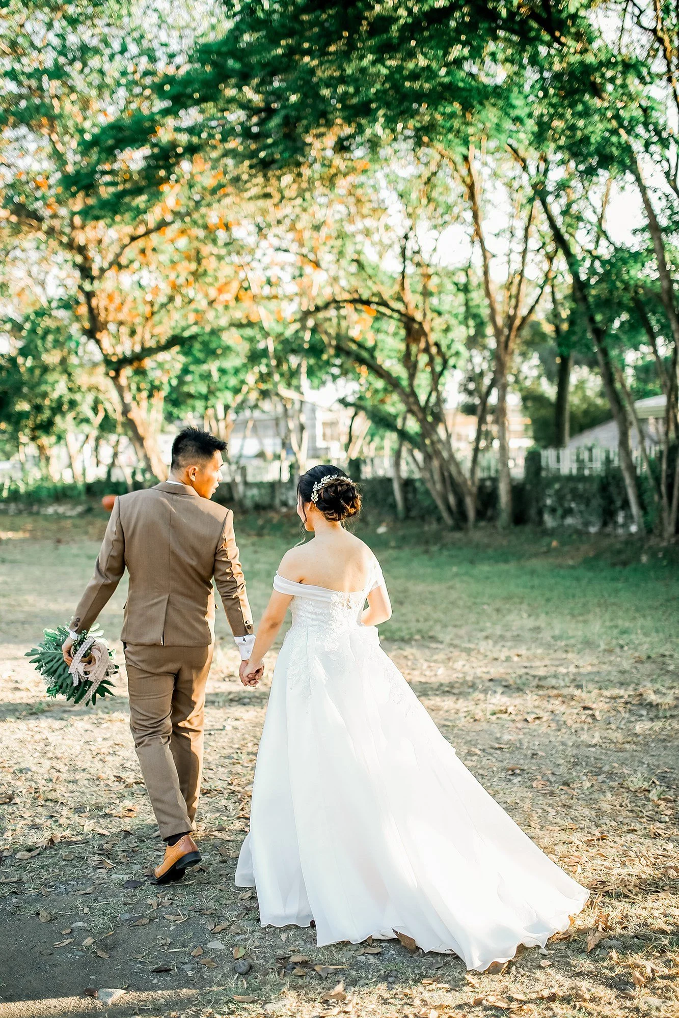 A newlywed couple walking hand in hand outdoors, with the bride in a white wedding gown and the groom in a beige suit, under a canopy of green trees with sunlight filtering through.