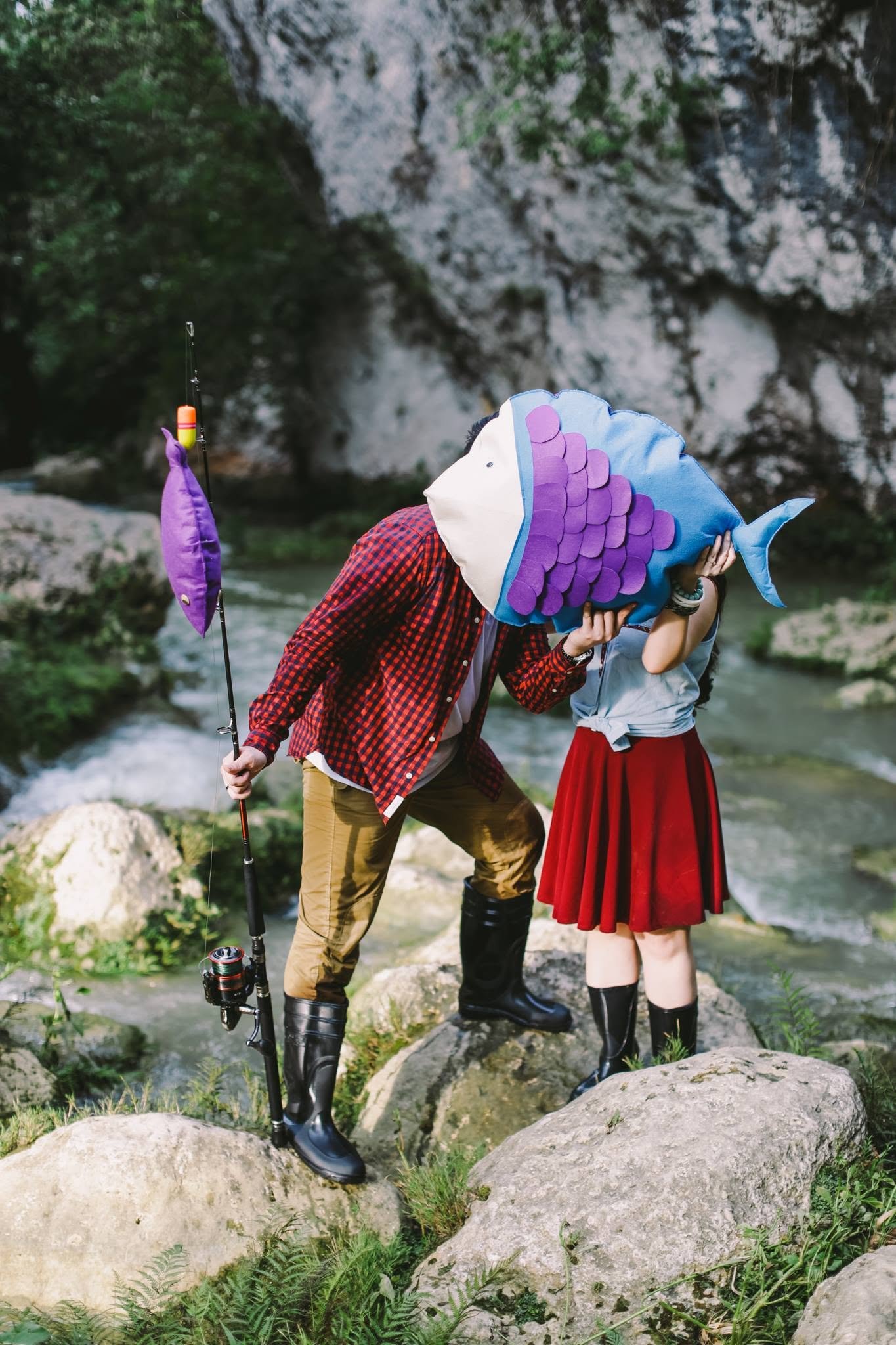Two people with fish-shaped head coverings standing by a river in a rocky, green outdoor setting, one holding a fishing rod.