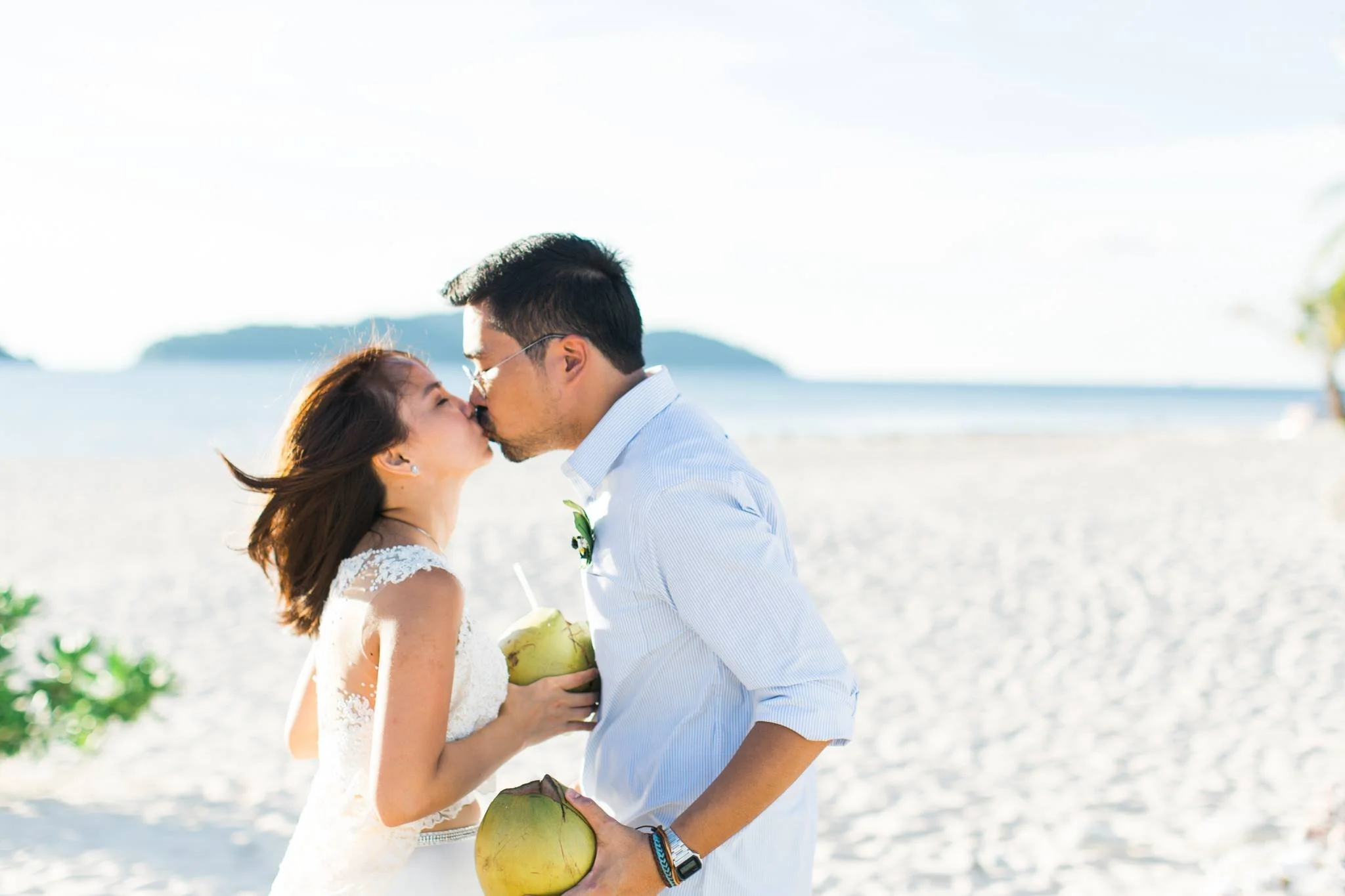 A couple sharing a kiss on the beach during the daytime, with the woman holding a coconut and the man wearing sunglasses and a light blue shirt.