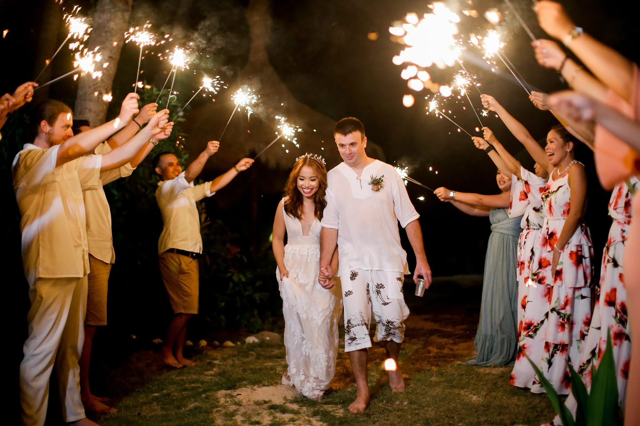 A newlywed couple walking hand in hand among friends holding sparklers at night during a celebration or wedding reception.