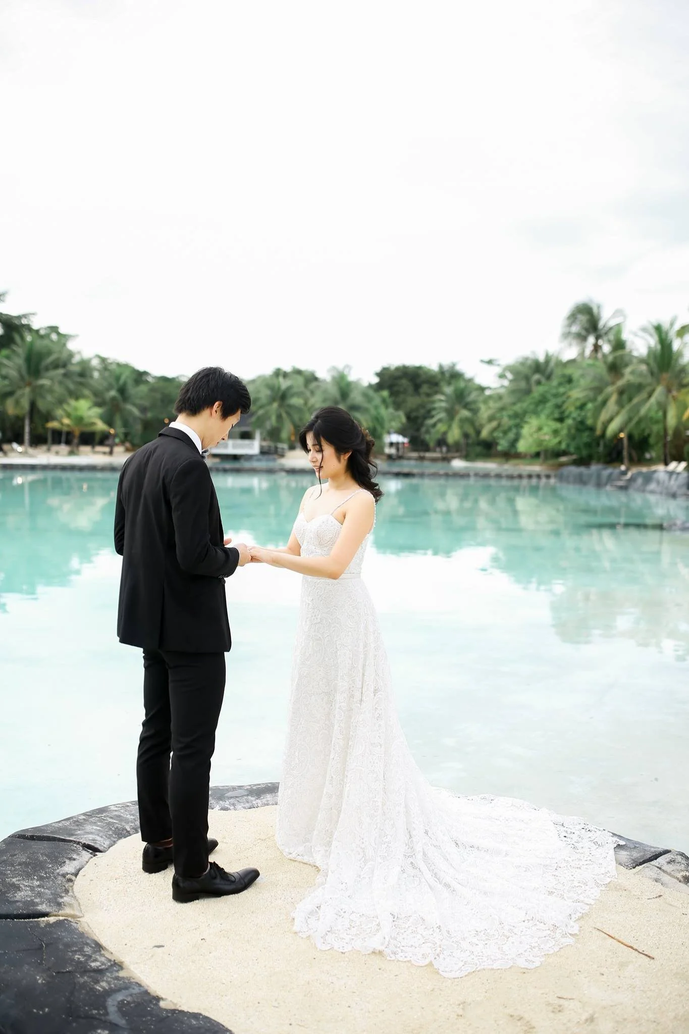 A bride and groom standing on a rocky and sandy area near a body of water, holding hands and facing each other during a wedding ceremony, with trees and a cloudy sky in the background.