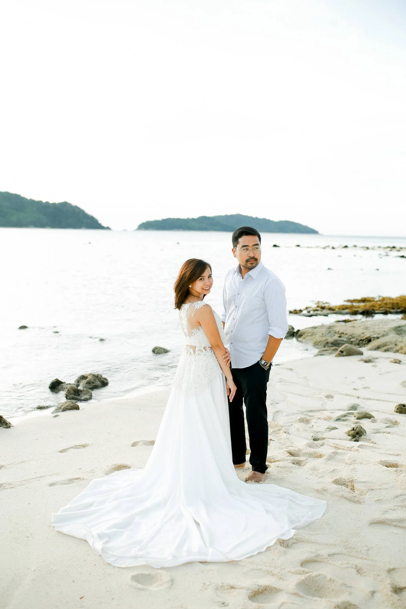A bride in a white wedding dress and a groom in a light blue shirt and black pants standing on a sandy beach near rocks, with water and landmasses in the background.