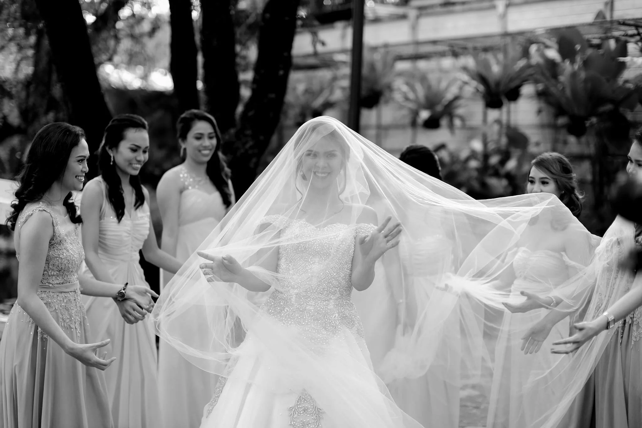 A bride in a wedding gown smiling, surrounded by bridesmaids holding up her veil outdoors.