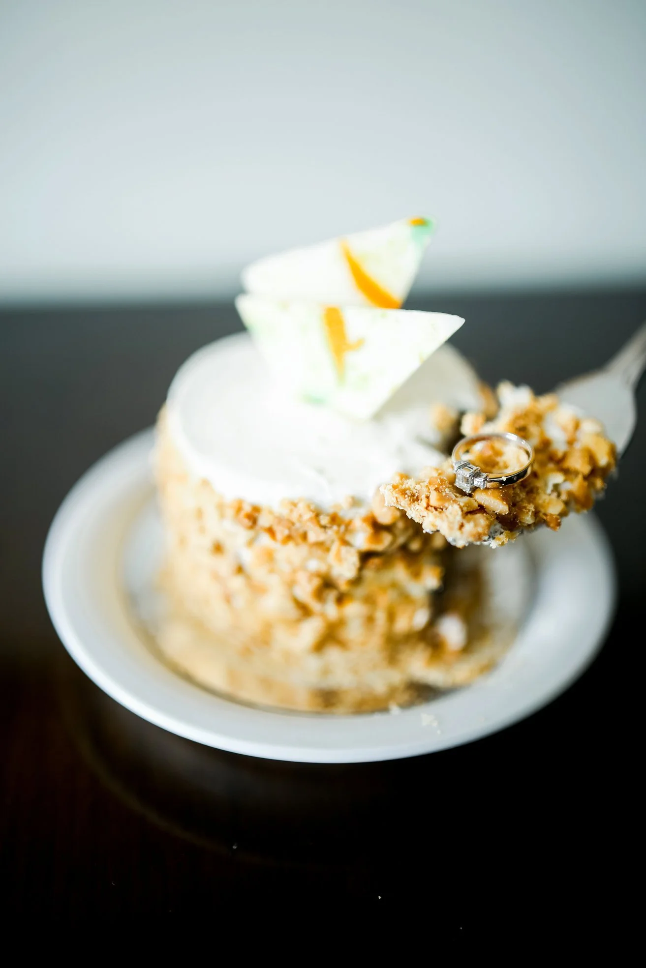 A slice of cake with white frosting topped with two rectangular slices of green and orange jelly-like candy, a ring with a diamond, and chopped nuts on the edge, served in a white bowl.