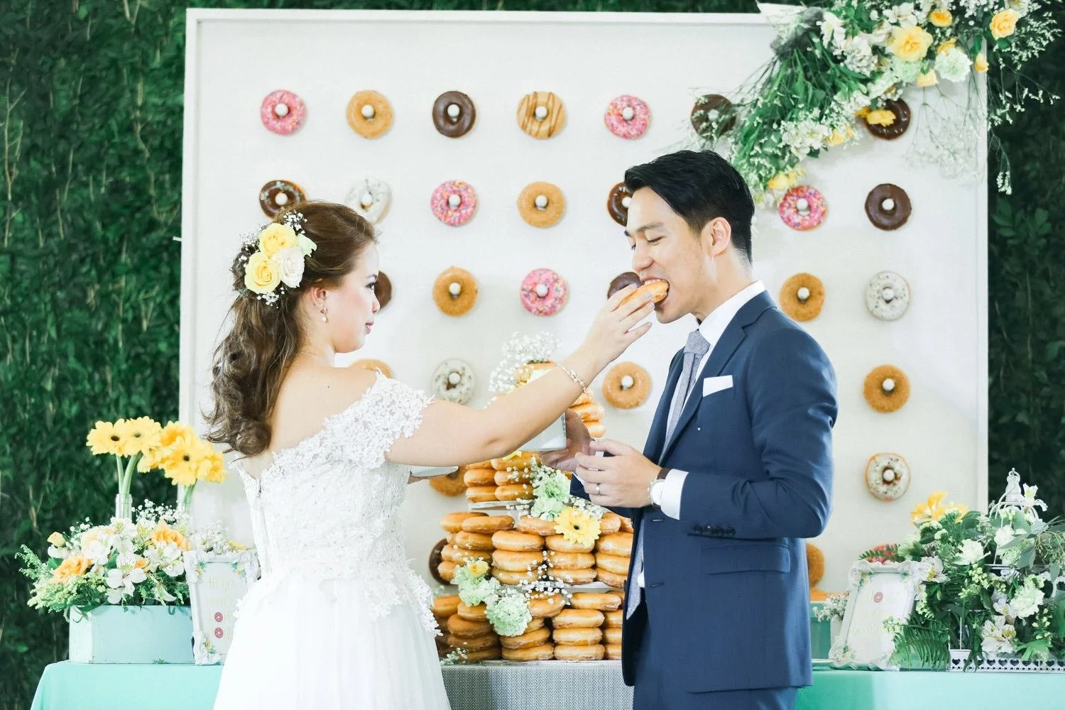 A bride in a white wedding dress feeds a man in a navy suit a donut during a wedding celebration, with a decorated donut wall and floral arrangements in the background.