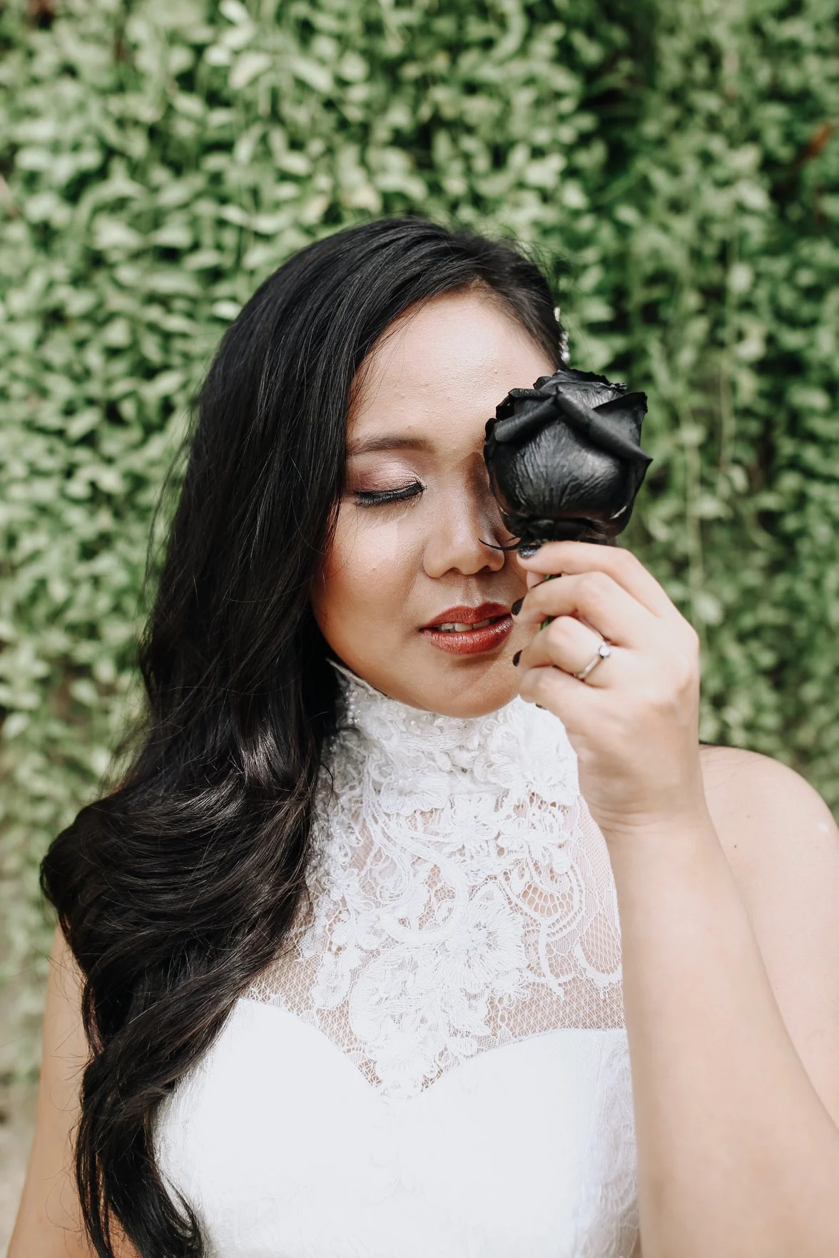 A woman with long black hair and light skin holding a black rose in front of her eye, wearing a white lace top, standing outdoors in front of green foliage.