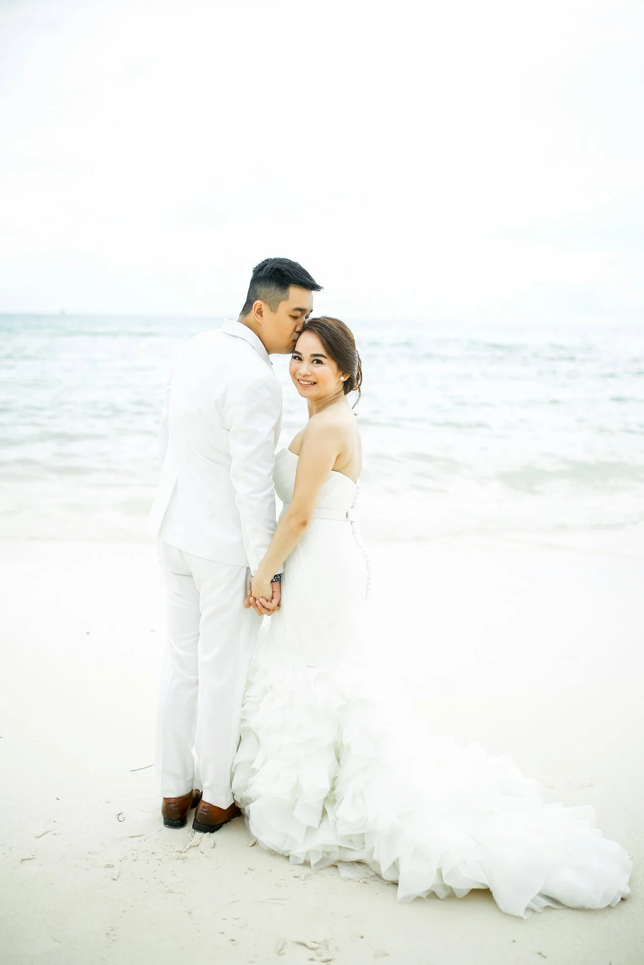 A bride and groom stand on a beach, holding hands. The groom is kissing the bride on the forehead, and they are both smiling. The bride is wearing a white strapless wedding gown, and the groom is dressed in a white suit with brown shoes. The backgrou